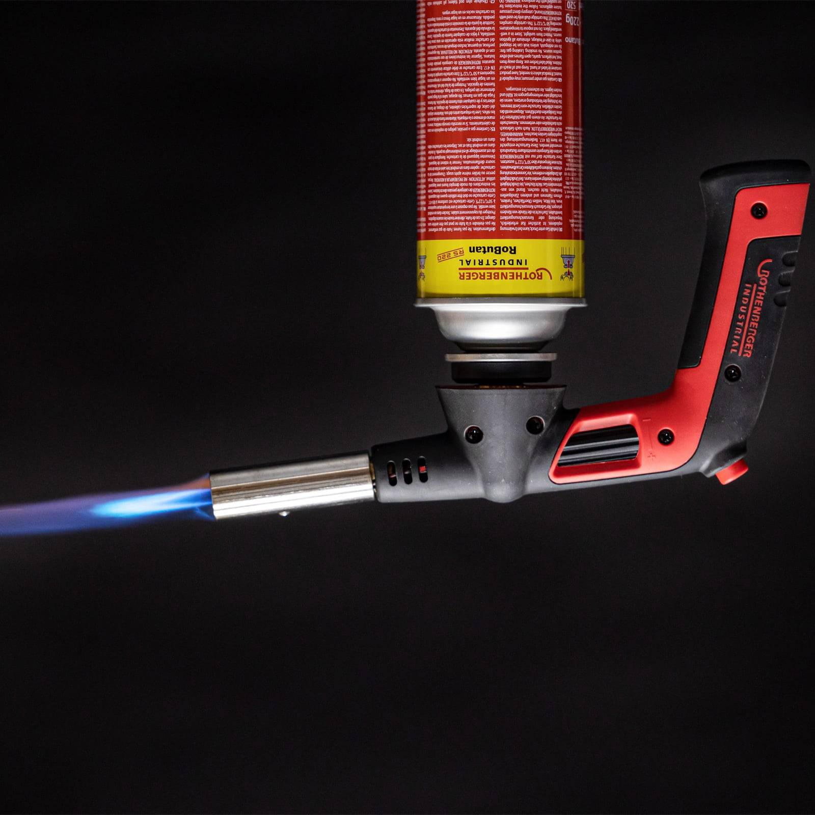 Eine tragbare Propangasfackel wirft eine blaue Flamme und zeigt ihre Verwendung für Heiz- oder Lötarbeiten. Die Gasflasche ist rot mit Text und Sicherheitswarnungen.