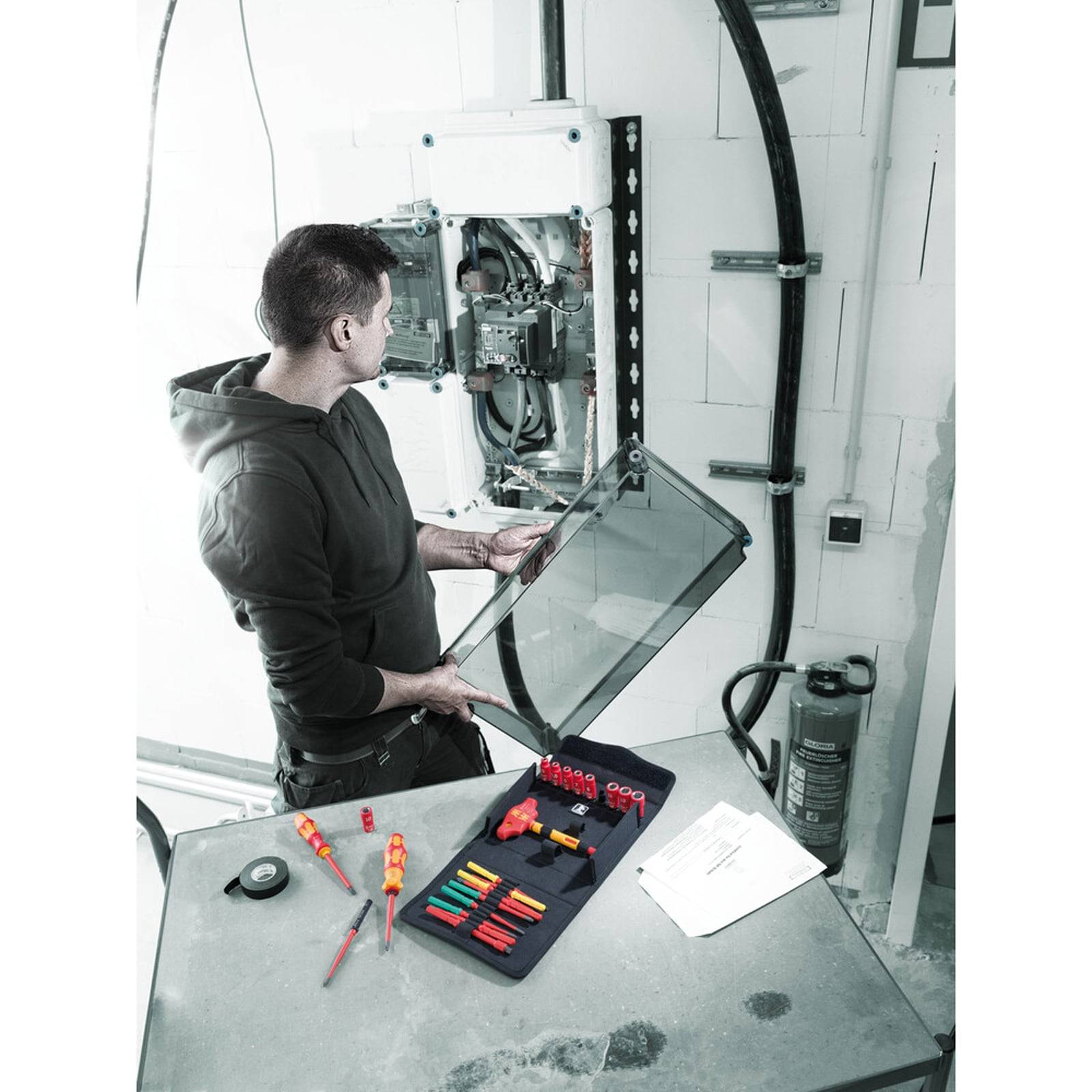 Elektriker, der an einer offenen elektrischen Schalttafel arbeitet, mit verschiedenen Werkzeugen auf einem nahegelegenen Tisch. Ein Feuerlöscher befindet sich im Hintergrund.