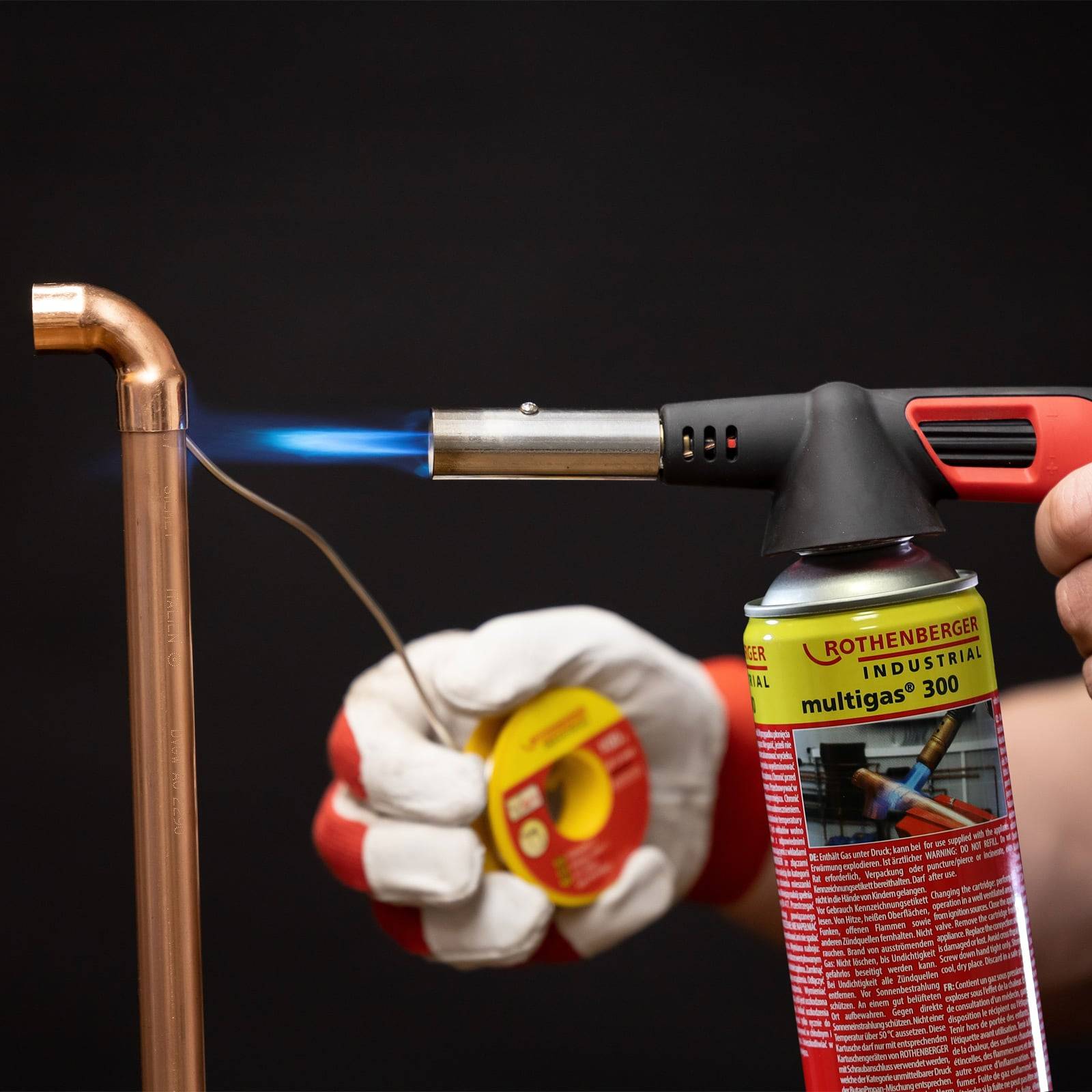 Eine Person verwendet eine Fackel und Flamme zum Löten eines Kupferrohrs und hält dabei eine Rolle Lötzinn in der anderen Hand, wobei sie Sicherheitshandschuhe trägt.