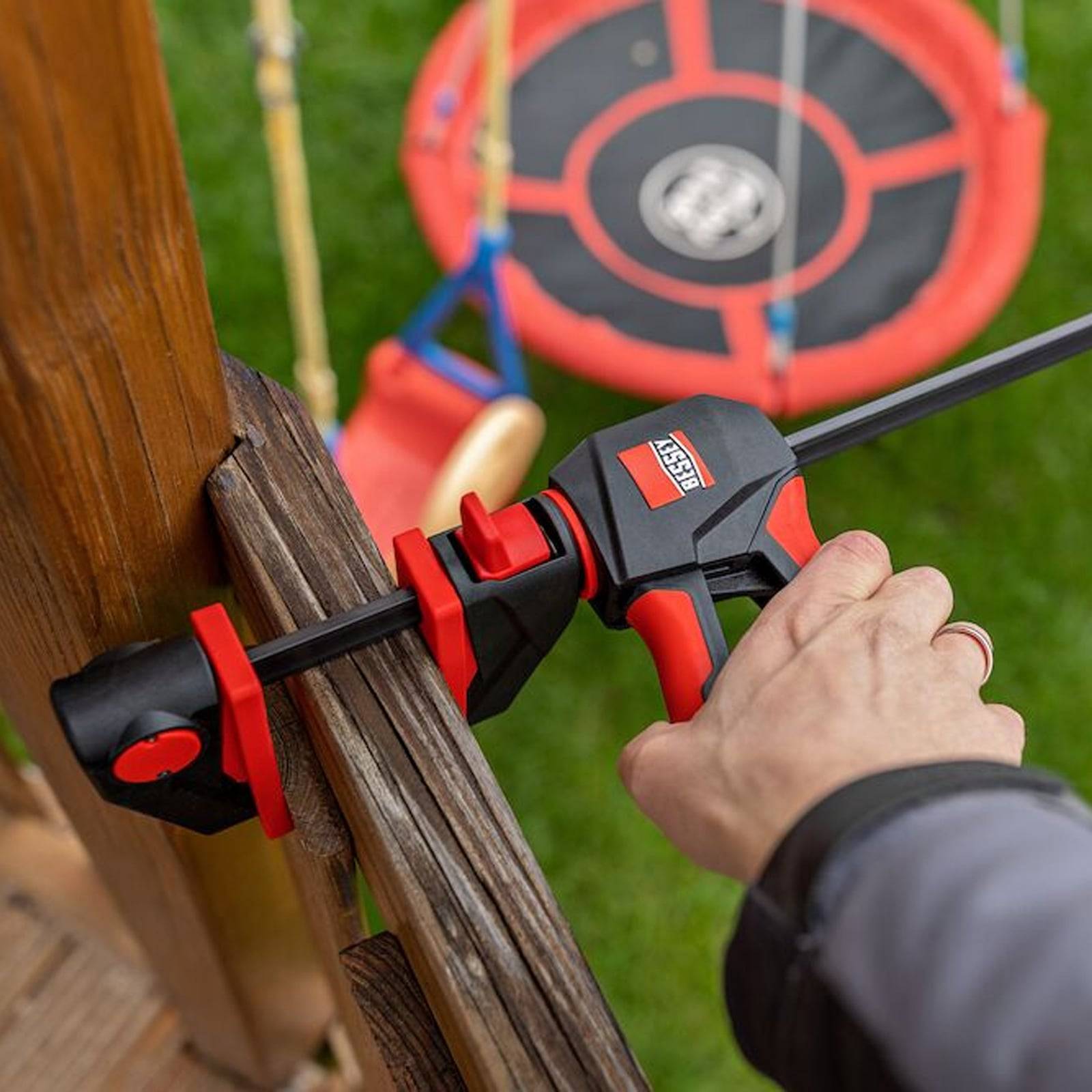 Eine Person verwendet eine Schnellspannklemme, um einen Holzpfosten zu befestigen, während im unscharfen Hintergrund eine Schaukel auf einem Rasenbereich zu sehen ist.
