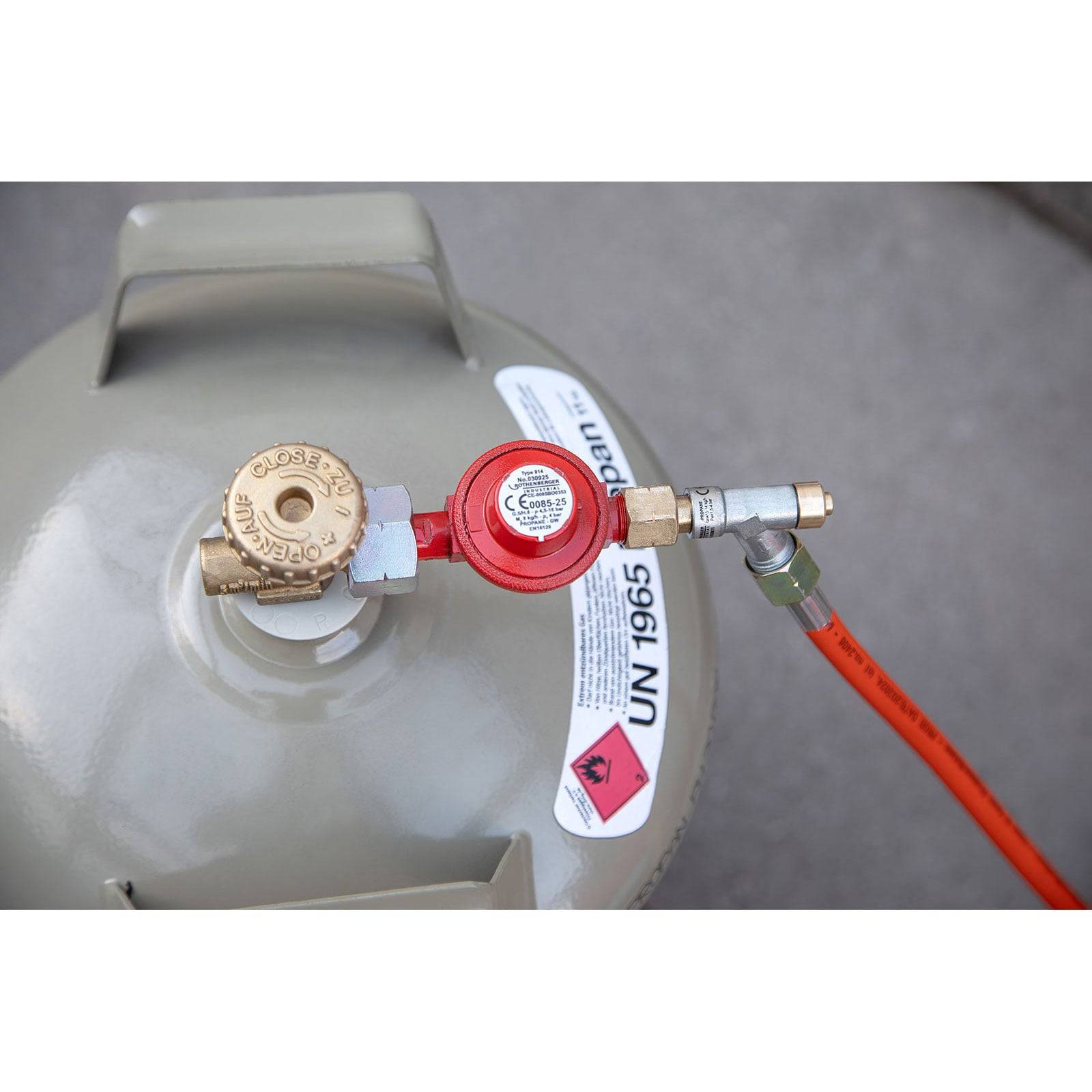 Draufsicht eines Gaszylinders mit Ventil und rotem Druckregler, der an einen orangefarbenen Schlauch angeschlossen ist. Sicherheitsetikett mit der Aufschrift 