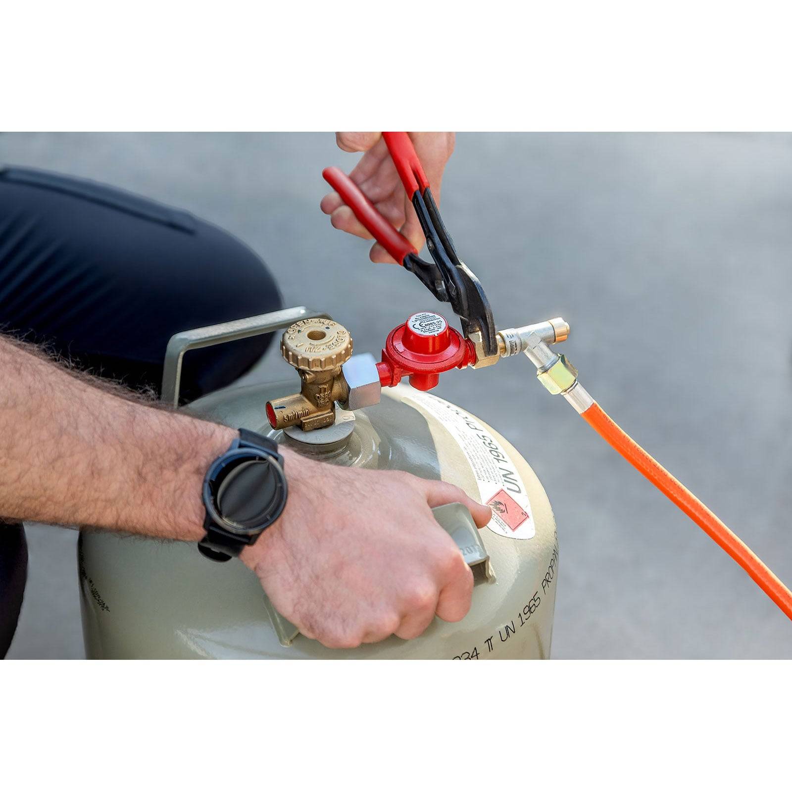 Eine Person mit einer schwarzen Armbanduhr benutzt eine Zange, um ein Ventil an einem Propangaszylinder zu befestigen, das mit einem orangefarbenen Schlauch verbunden ist.