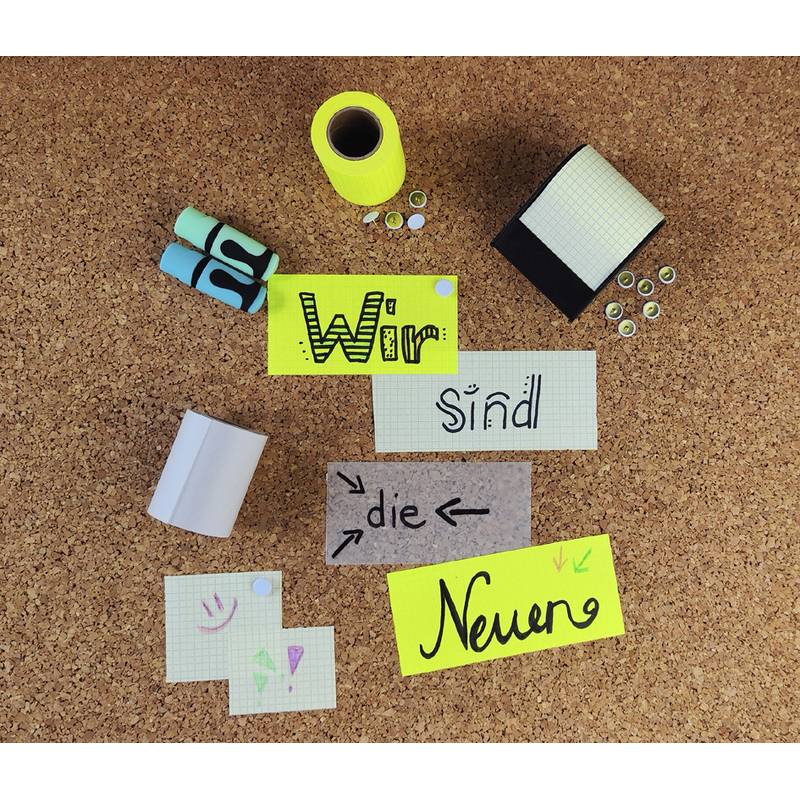 Eine Pinnwand zeigt die deutsche Phrase 'Wir sind die Neuen', geschrieben auf farbigem Papier, umgeben von Büromaterialien.