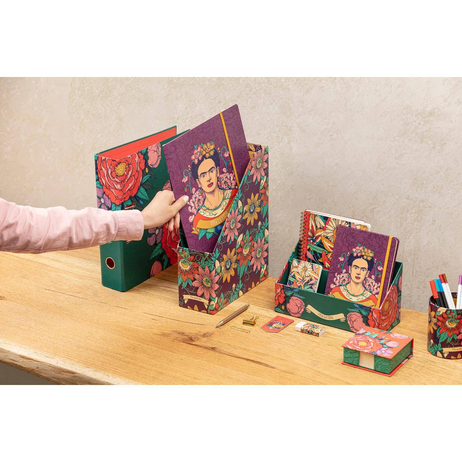 Eine Person organisiert eine Sammlung von Frida-Kahlo-Motiv Büromaterialien, einschließlich Aktenhaltern, Notizbüchern und Stiftehaltern, auf einem Holzschreibtisch.