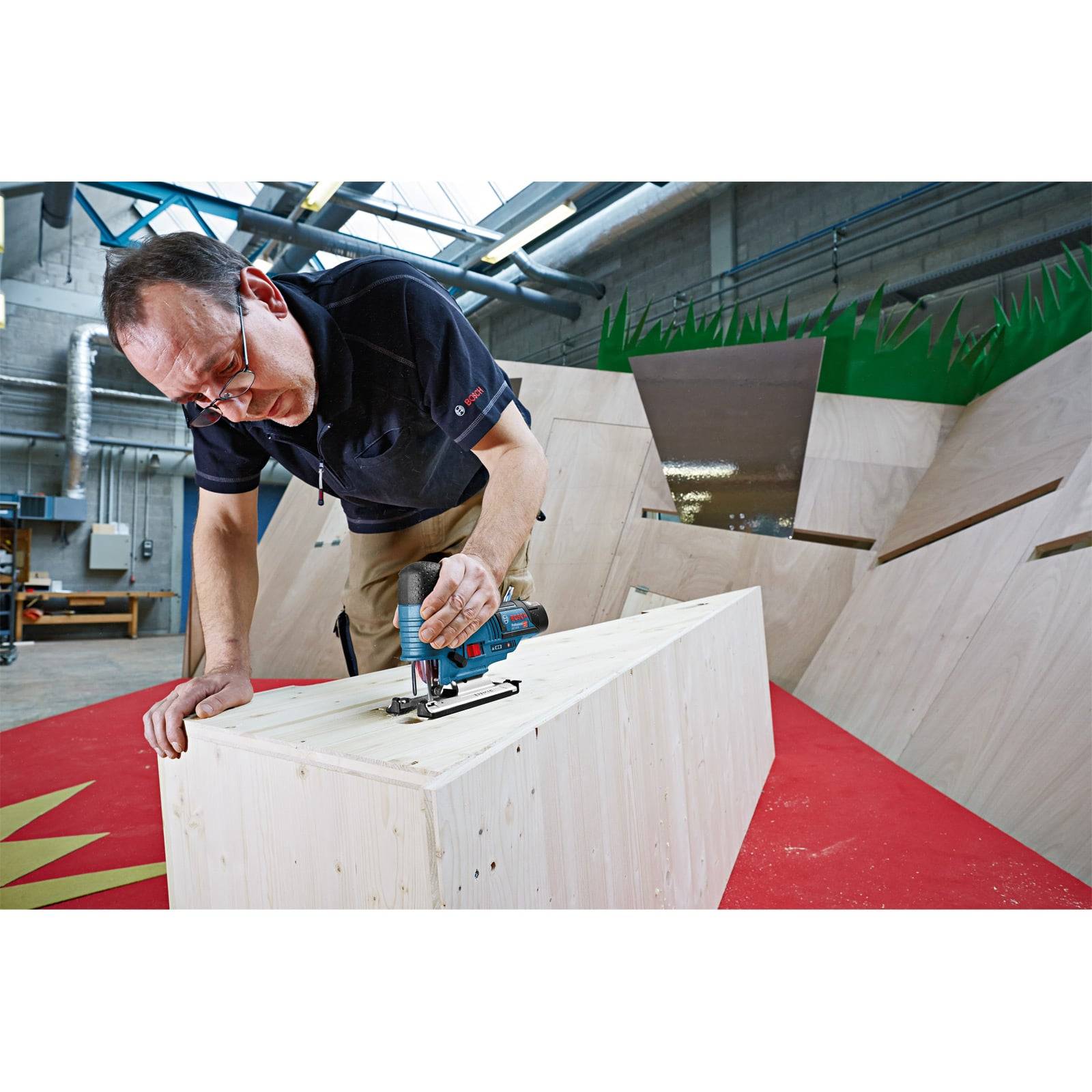 Eine Person verwendet eine elektrische Stichsäge, um in einer Werkstatt ein Stück Holz zu schneiden. Die Umgebung ist industriell mit Holzstrukturen und rotem Fußboden.