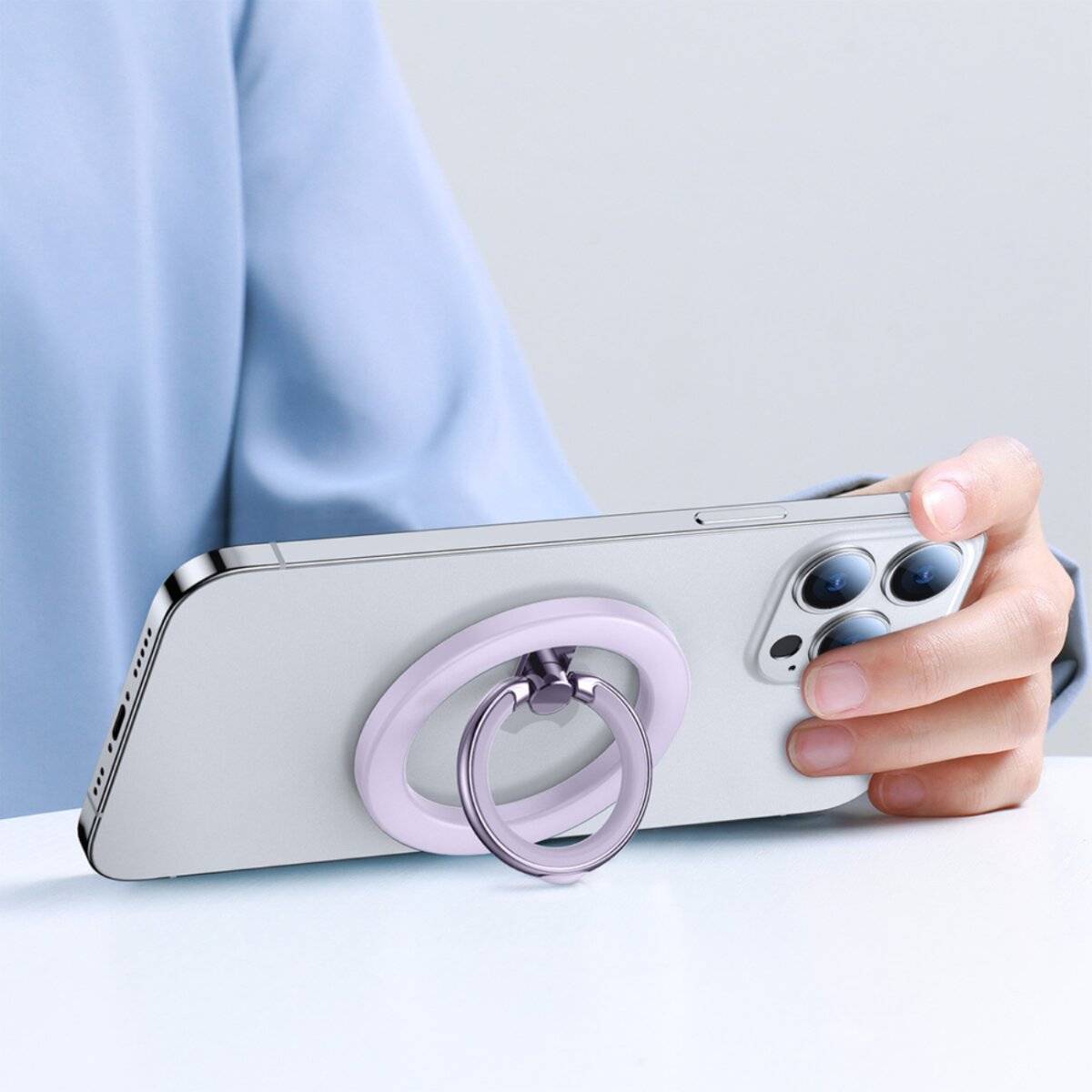 Eine Person hält ein Smartphone mit einem lila Ringhalter auf der Rückseite und nutzt diesen, um das Telefon auf einer hellblauen Oberfläche in einem Winkel abzustützen.