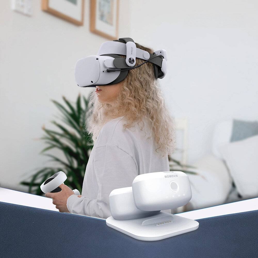 Eine Person trägt ein VR-Headset und sitzt in einem hellen Raum mit Pflanzen und gerahmten Bildern. Ein kabelloses Ladegerät und VR-Controller liegen auf dem Tisch.