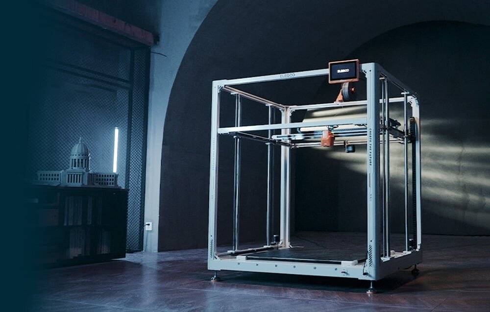 Ein großer 3D-Drucker in einem dunkel beleuchteten Raum, der aktiv ein Objekt druckt. Ein Bildschirm zeigt den Druckerstatus an. Die Umgebung ist industriell und modern.