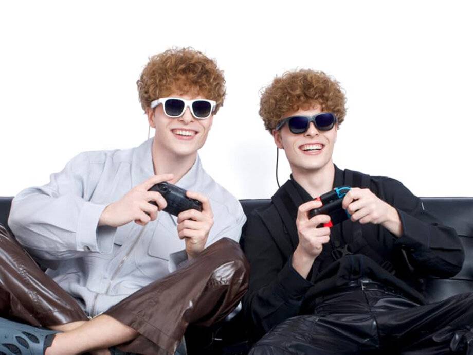 Zwei lächelnde Personen, die Sonnenbrillen tragen, spielen Videospiele mit Controllern, sitzen sich eng beieinander gegenüber vor einem einfachen weißen Hintergrund.