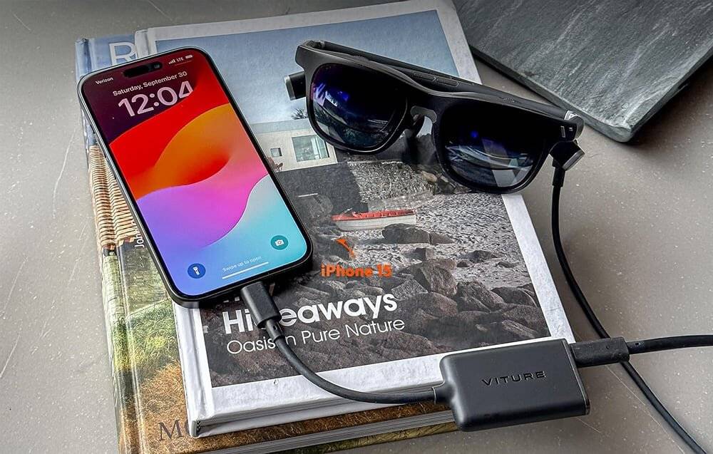 Ein Smartphone mit einem farbenfrohen Bildschirm zeigt die Zeit und das Datum an und ist über ein Kabel mit einem Paar schwarzer Smartglasses auf einem Buch verbunden.
