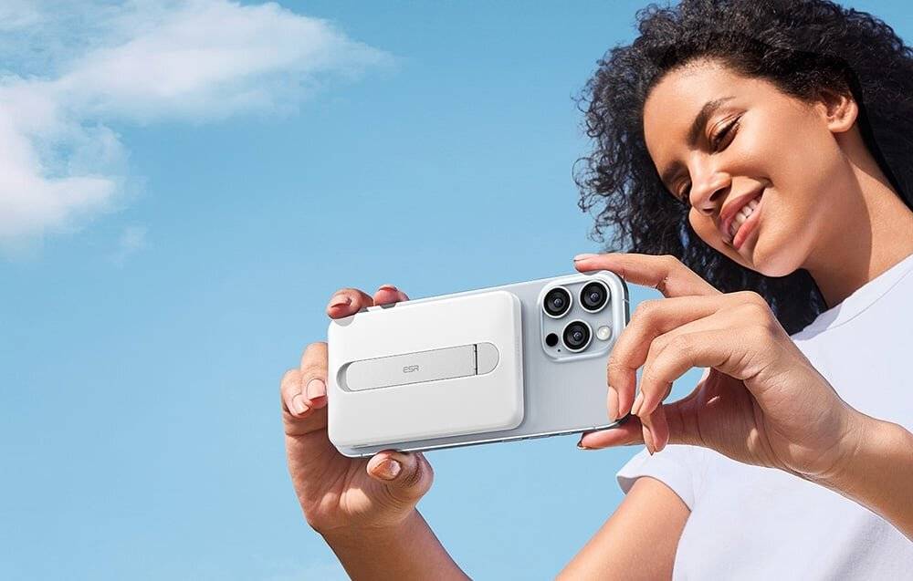 Eine Person hält ein Smartphone mit weißem Gehäuse und Zubehör gegen einen klaren blauen Himmel, lächelt sanft mit geschlossenen Augen.