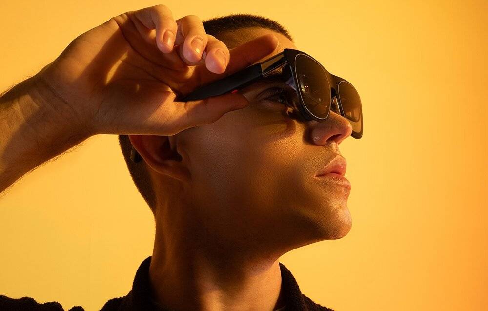 Eine Person mit Sonnenbrille blickt gegen einen gelben Hintergrund nach oben, während eine Hand auf ihrer Stirn ruht und sie nachdenklich wirkt.