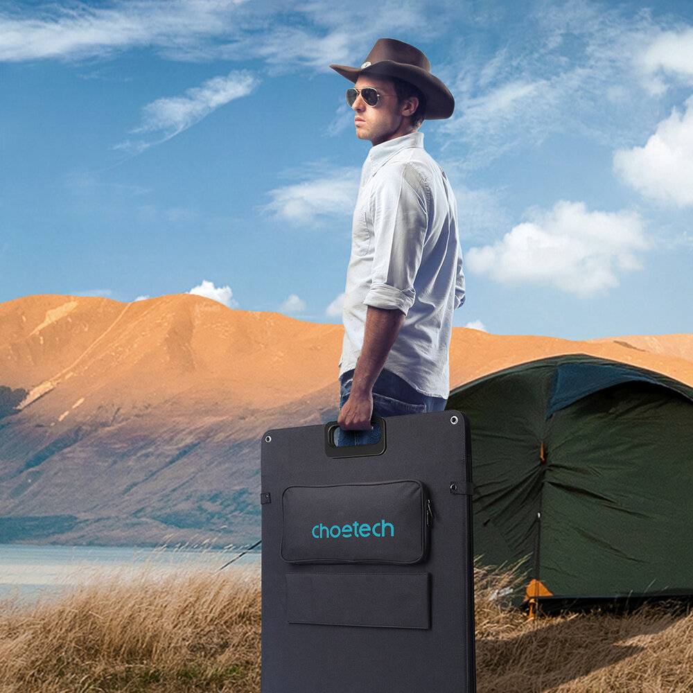 Eine Person mit einem Hut steht in der Nähe eines tragbaren Solarpanels und eines Zelts an einem See mit Bergen im Hintergrund unter einem bewölkten Himmel.