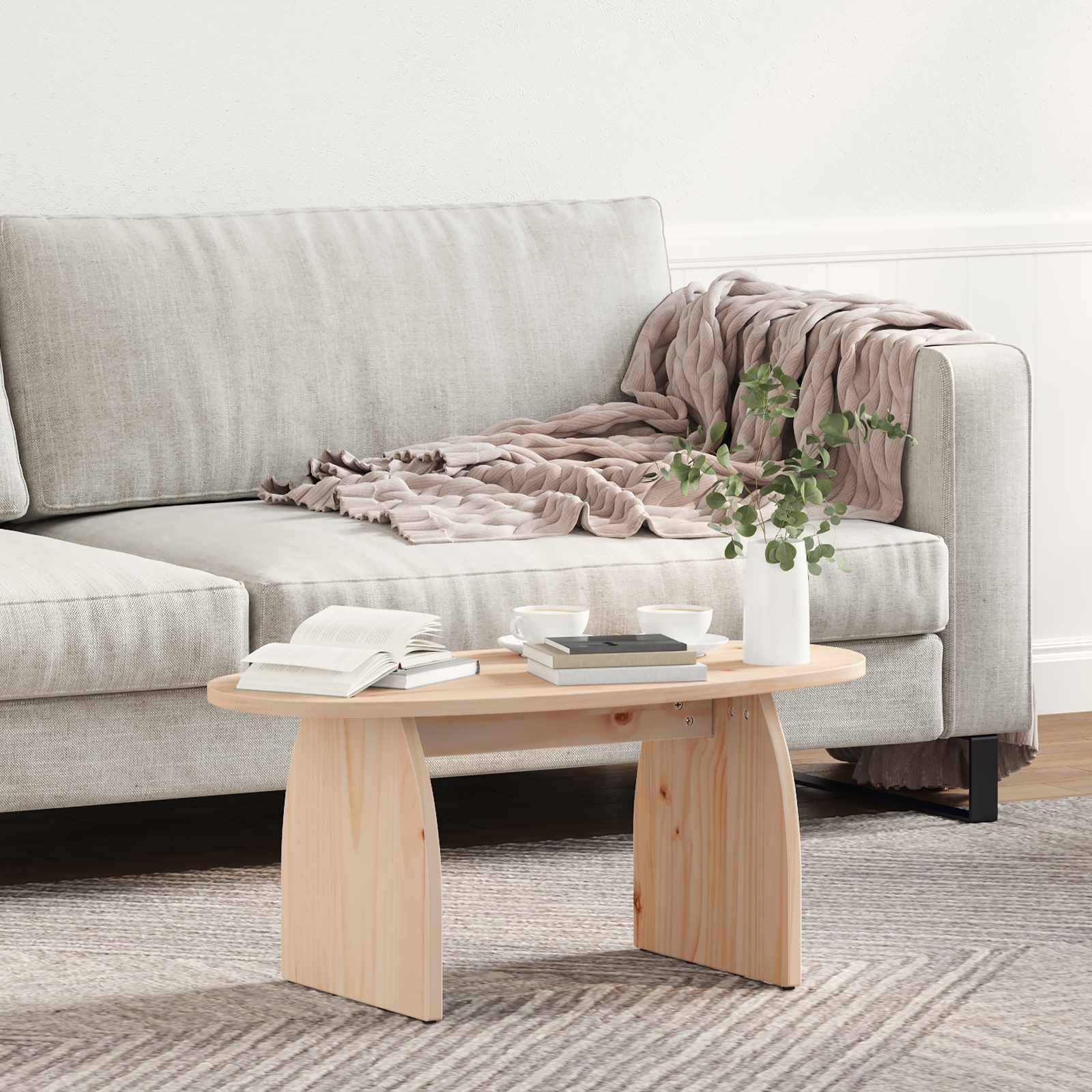Ein hellgrauer Sofa mit einer beigen Decke und Kissen, neben einem hellen Holzkaffeetisch. Auf dem Tisch stehen Bücher, Tassen und eine kleine Pflanze.