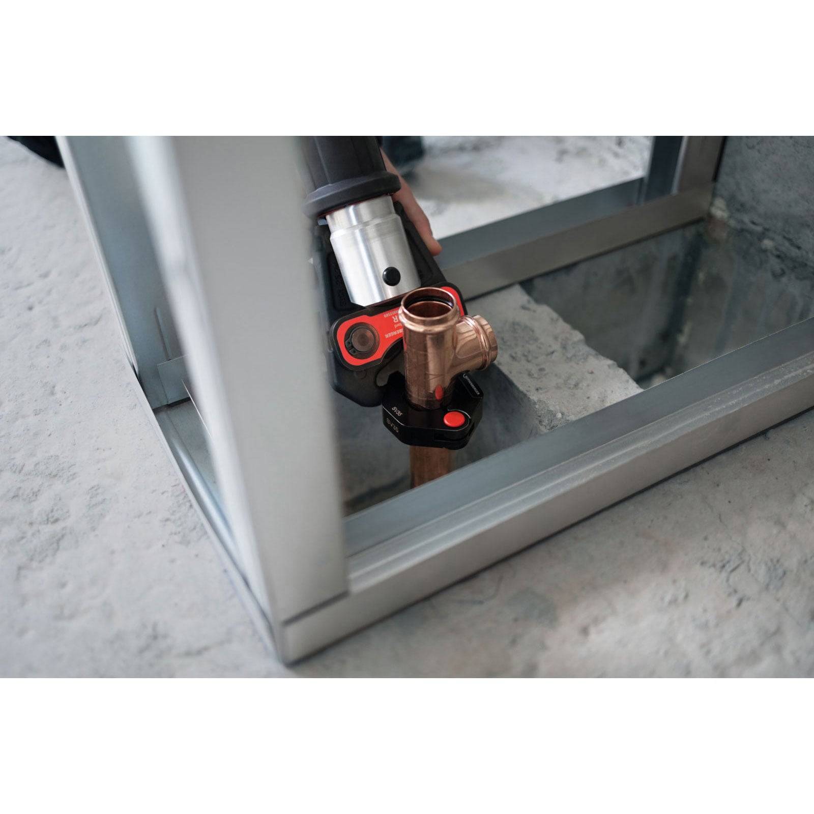 Eine Nahaufnahme einer Person, die ein Rohrpressgerät verwendet, um Kupferrohre auf einer reflektierenden Oberfläche zu verbinden, gerahmt von einer Metallstruktur.