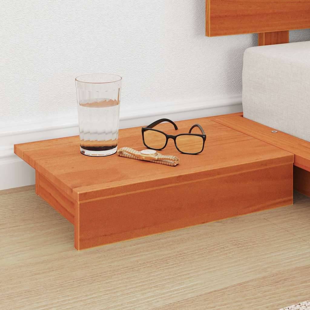 Ein Nachttisch aus Holz mit einem Wasserglas, einer Brille und einem Buch darauf, neben einem Bett mit hellem Bodenbezug.