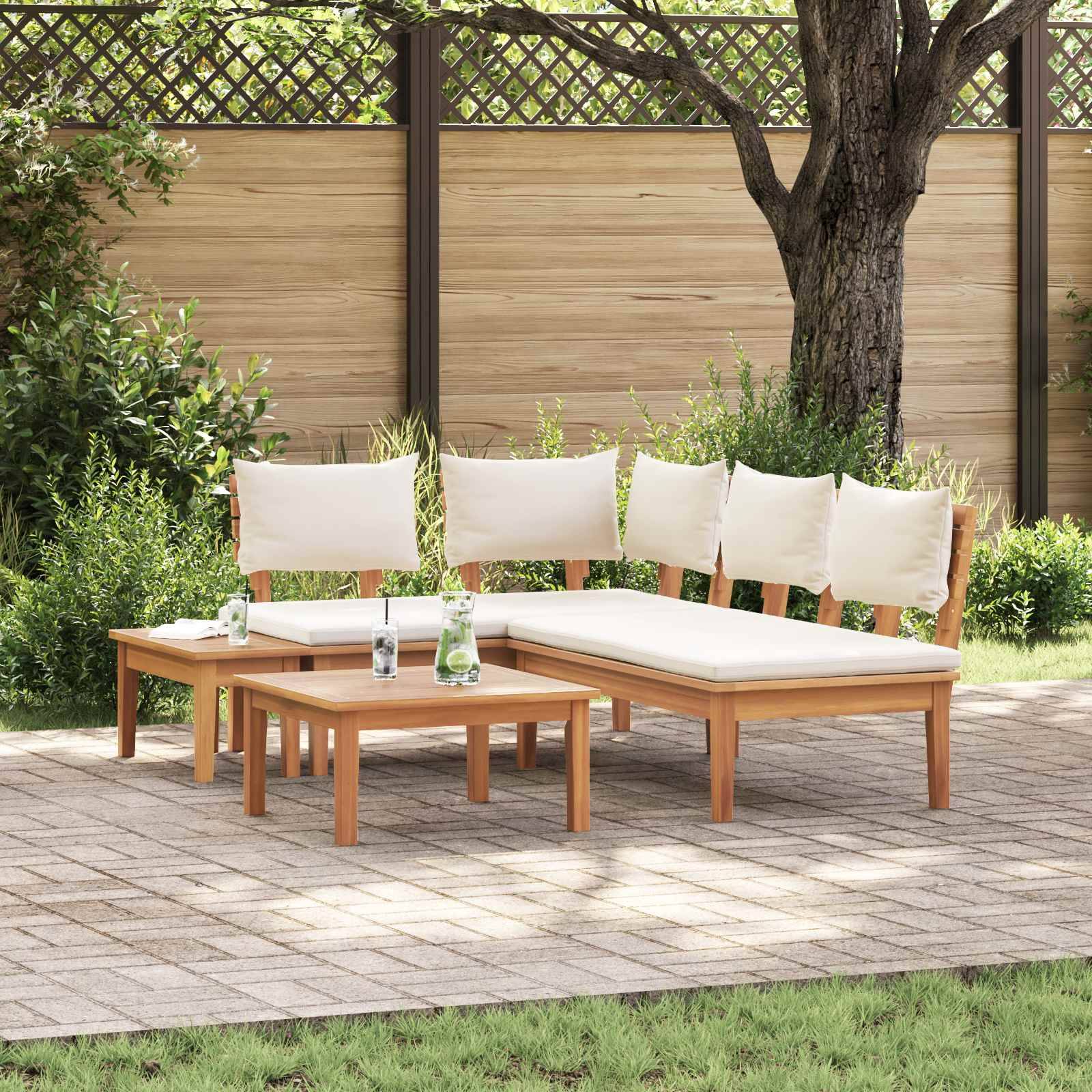 Eine Lounge-Sitzgruppe aus Holz mit weißen Kissen steht auf einer Steinterrasse neben Grünpflanzen und einem großen Baum.