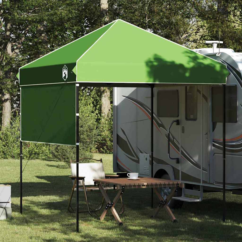 Ein grünes Blätterdach bedeckt einen kleinen Außenessbereich mit einem Tisch und zwei Stühlen in der Nähe eines geparkten Wohnmobils in einer bewaldeten Umgebung.