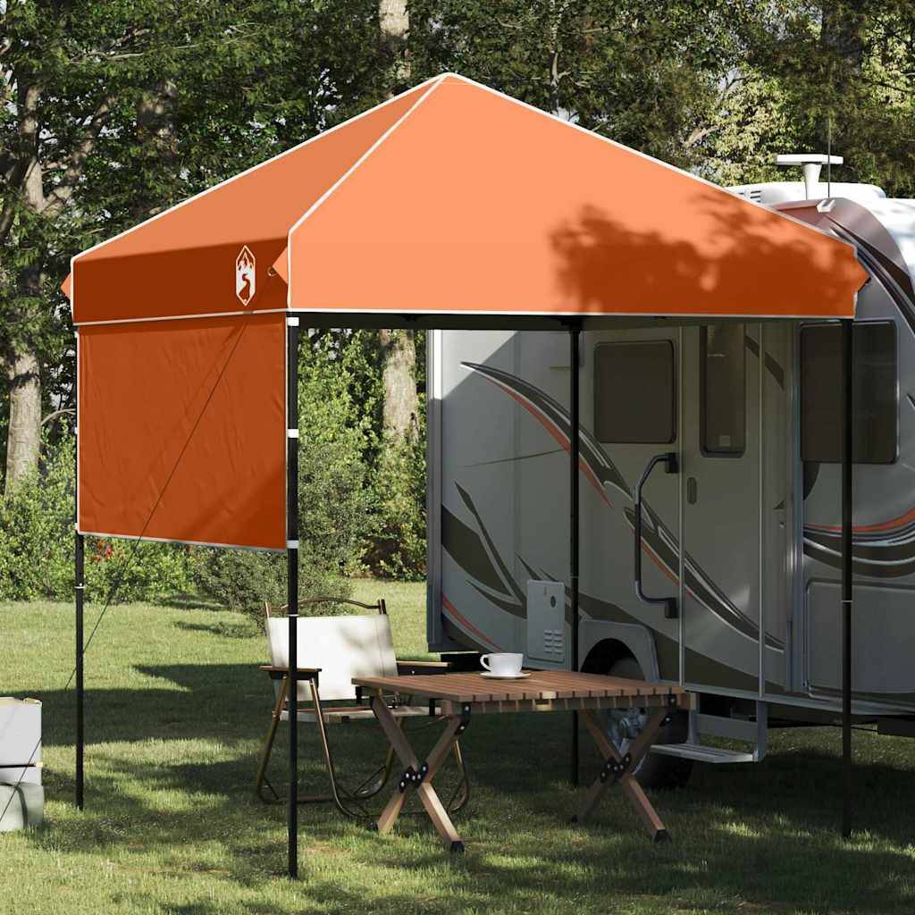 Ein orangefarbenes Pop-up-Zelt ist neben einem Wohnmobil aufgestellt, mit einem Holztisch und Stühlen darunter, umgeben von Bäumen.