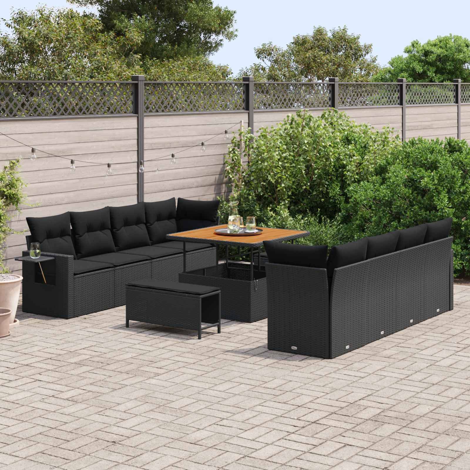 Outdoor-Sitzbereich mit schwarzen Korbmöbeln und Tischen auf einer gepflasterten Terrasse, umgeben von grünen Büschen und einem Holzzaun.