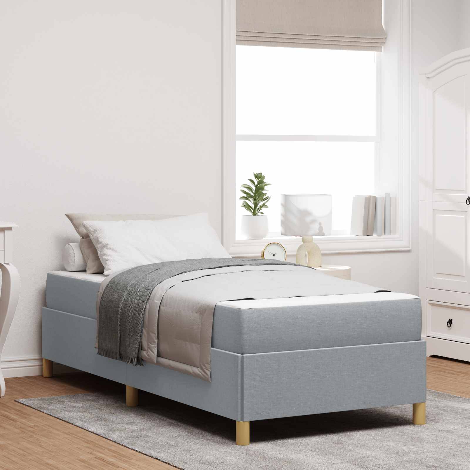 Minimalistisches Schlafzimmer mit einem Einzelbett, das mit grauer und weißer Bettwäsche bezogen ist, neben einem Fenster und einem kleinen Nachttisch in einem hellen, neutralen Raum.