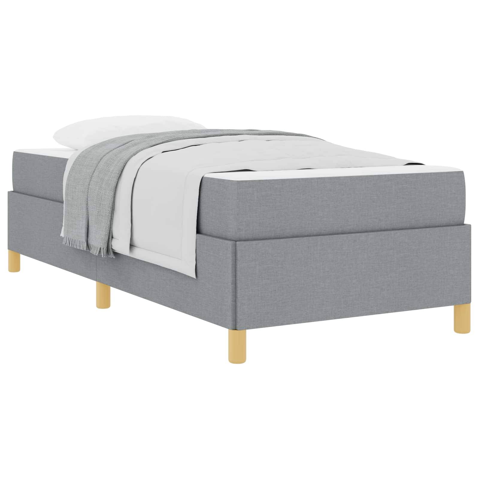 Ein einfaches Bett mit einem grauen Stoffrahmen und Holzbeinen, ausgestattet mit einer weißen Bettdecke, einem Laken und einer grauen Decke, die seitlich herunterhängt.