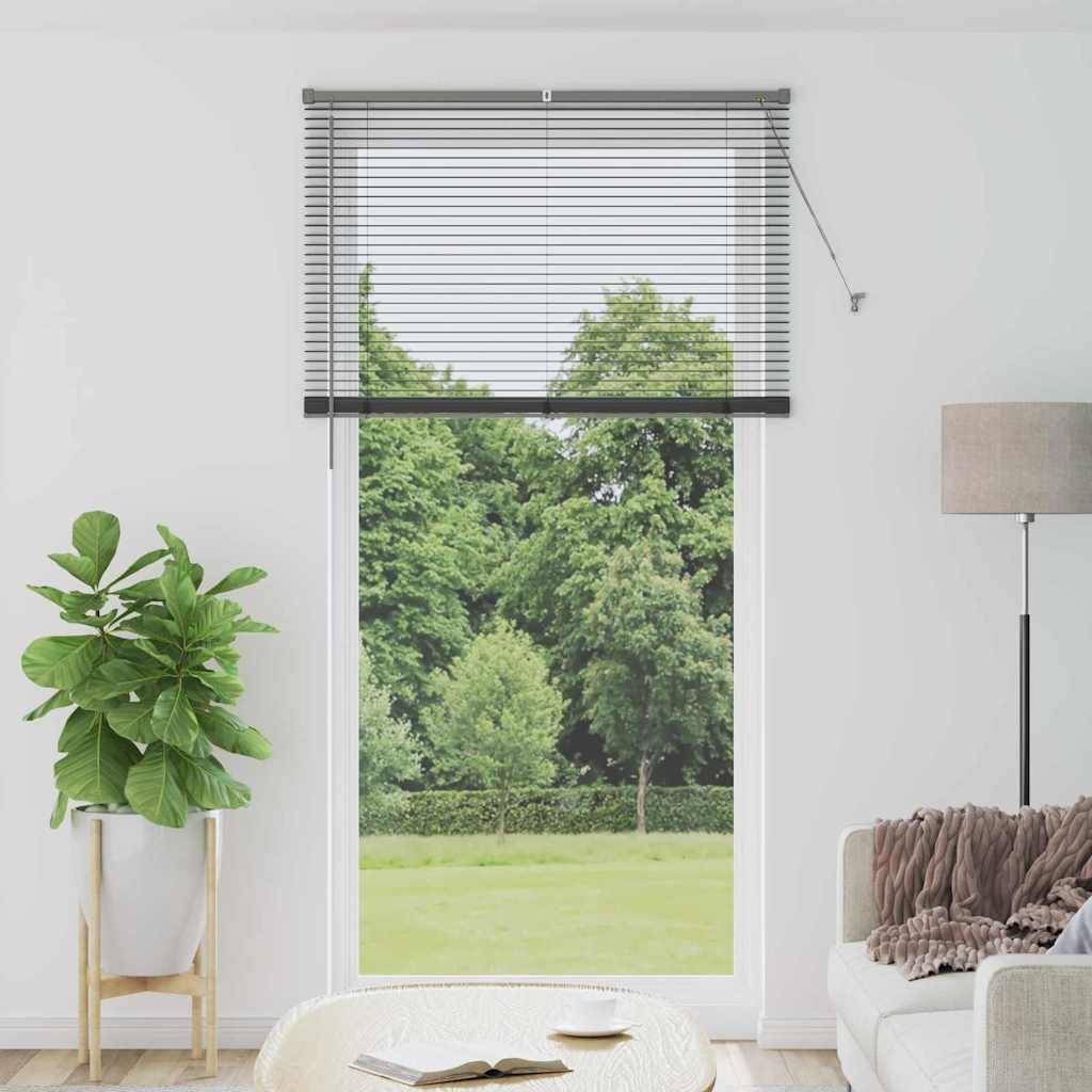 Ein großes Fenster mit horizontalen Jalousien, das den Blick auf üppig grüne Bäume draußen freigibt. Der Raum verfügt über eine Topfpflanze, ein graues Sofa, eine Lampe und einen Couchtisch.