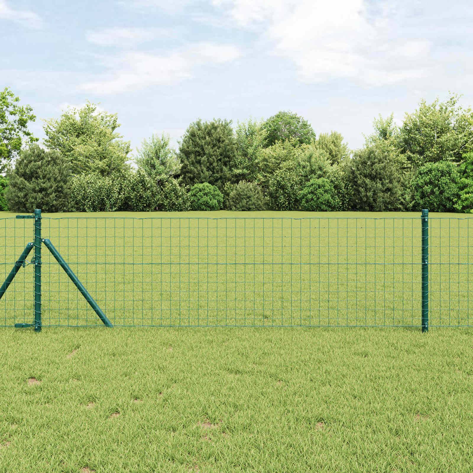 Ein grüner Metallzaun steht vor einem üppigen, grünen Feld mit dichten Bäumen und einem bewölkten Himmel im Hintergrund.