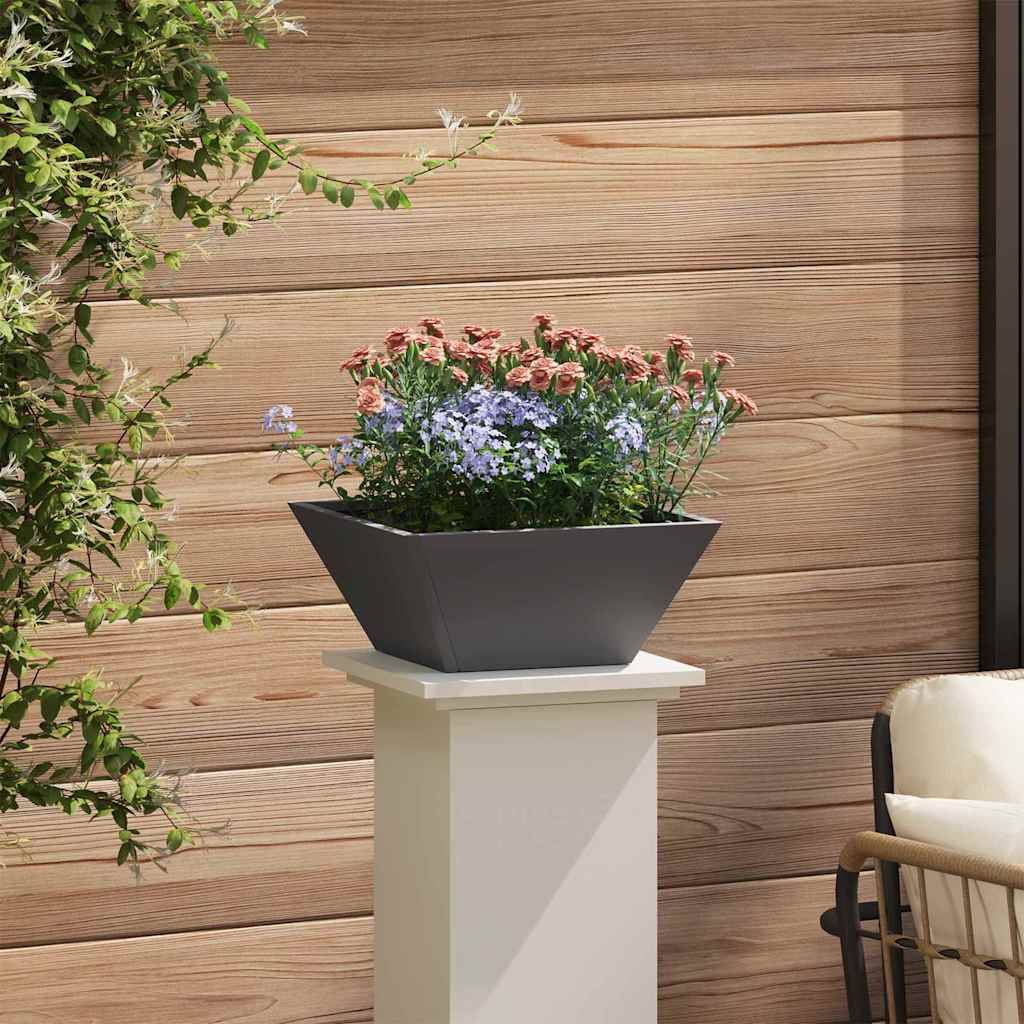 Ein dunkler, quadratischer Pflanzkasten mit rosa und violetten Blumen auf einem weißen Sockel, vor einer Holzwand mit Blattwerk auf der linken Seite.