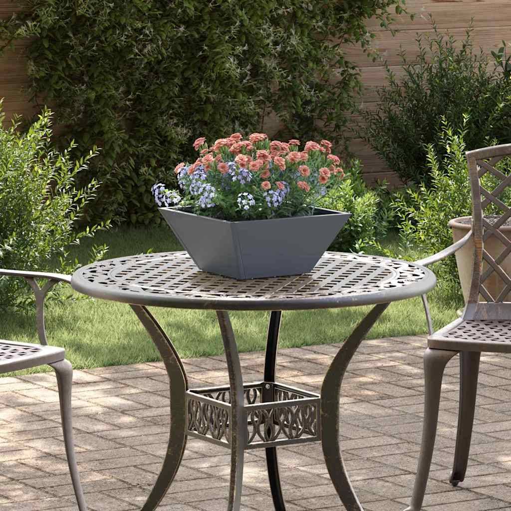 Ein runder Metalltisch für den Garten mit aufwendigem Design, auf dem ein quadratischer Pflanzentopf mit rosa und blauen Blumen steht, umgeben von Grünpflanzen.