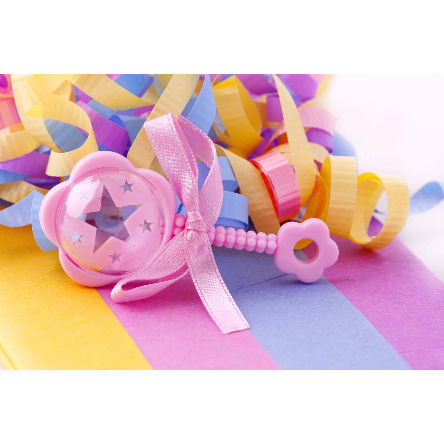 Rosa Babyklingel mit Band und Sternausschnitten liegt auf pastellfarbigem gestreiftem Papier zwischen bunten gekräuselten Bändern.