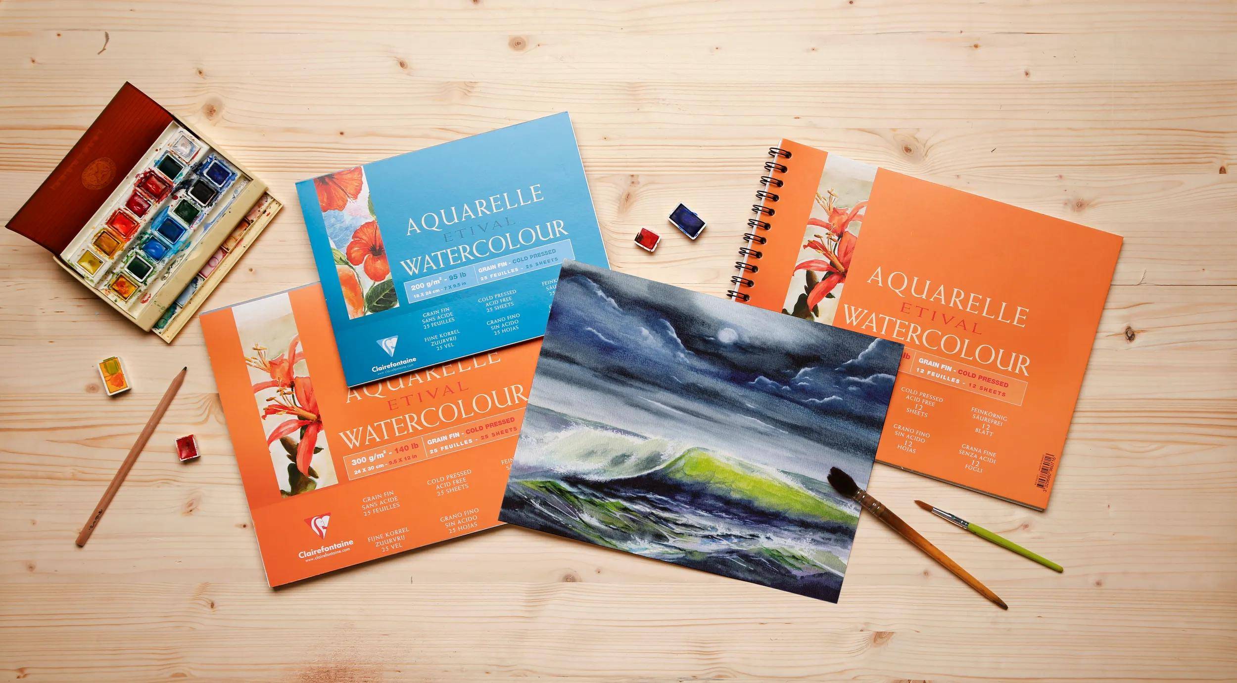 Aquarellmalstoffe auf einem Holzschreibtisch, einschließlich Malkasten, Pinsel, Skizzenbücher und Beispiel-Aquarellmalereien.