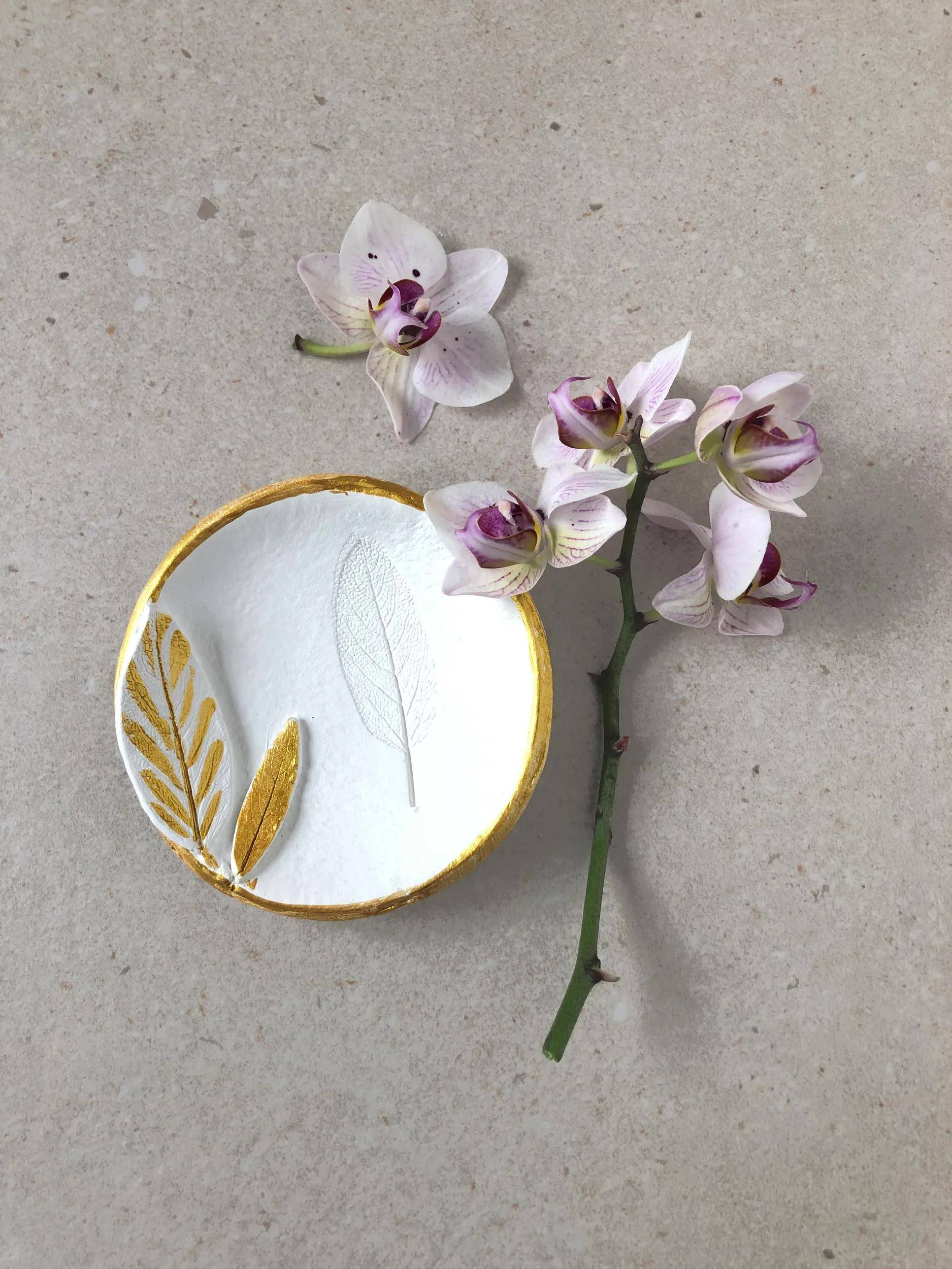 Ein kleines rundes weißes Schälchen mit Goldblatt-Verzierungen sitzt neben einem rosa Orchideen-Zweig auf einer grauen Oberfläche.