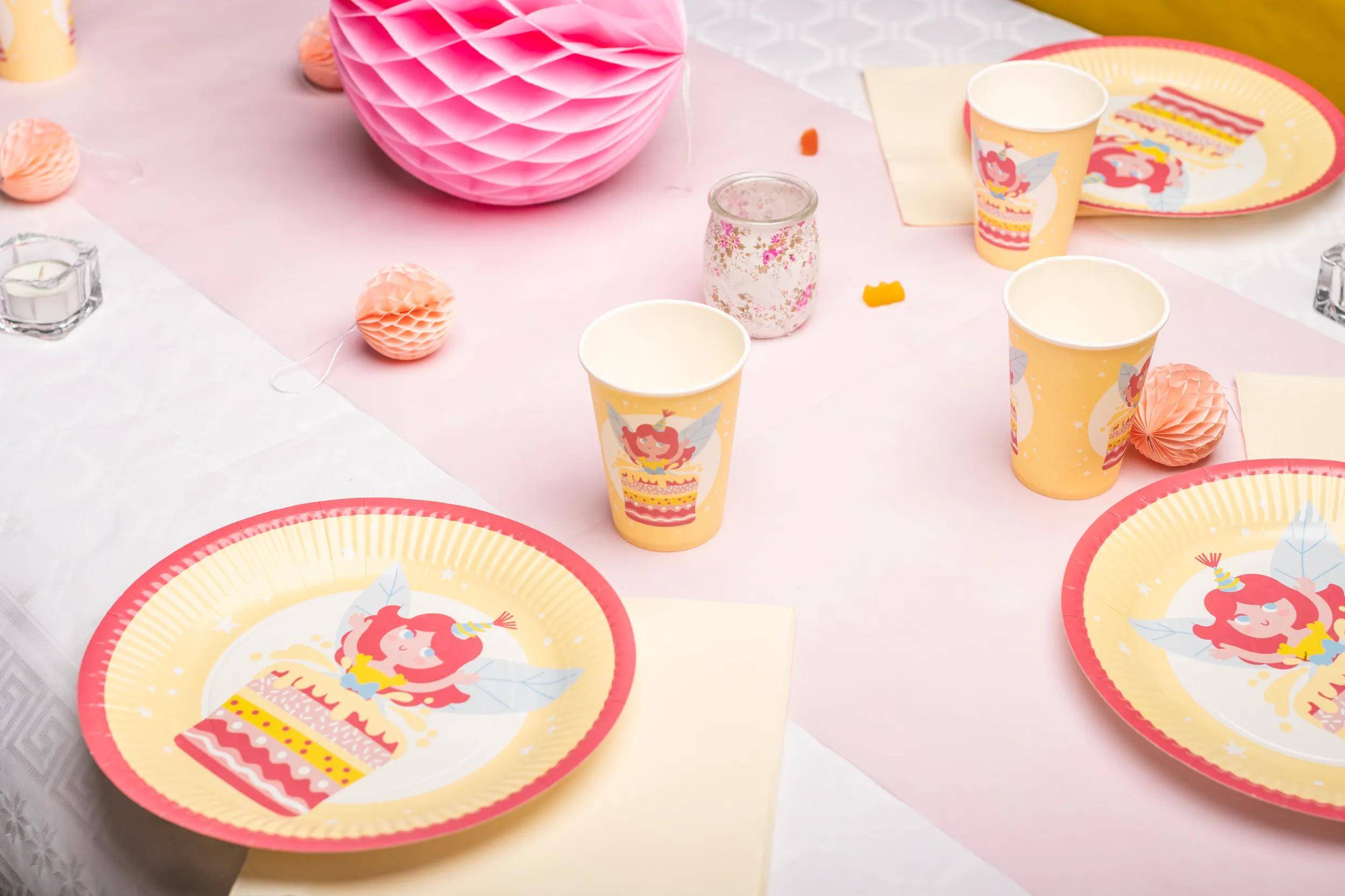 Eine Geburtstagsgedeck mit Partytellern und -bechern, die ein Mädchen mit einer Kuchendesign zeigen. Rosa und orangefarbene Dekorationen sind im Hintergrund.