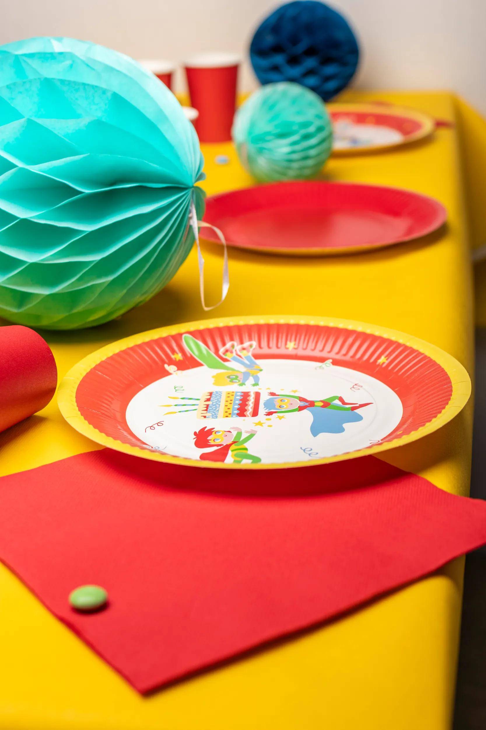 Ein Geburtstagstisch mit einer gelben Tischdecke, mit roten Papptellern und einer dekorativen blauen Papierlaterne.