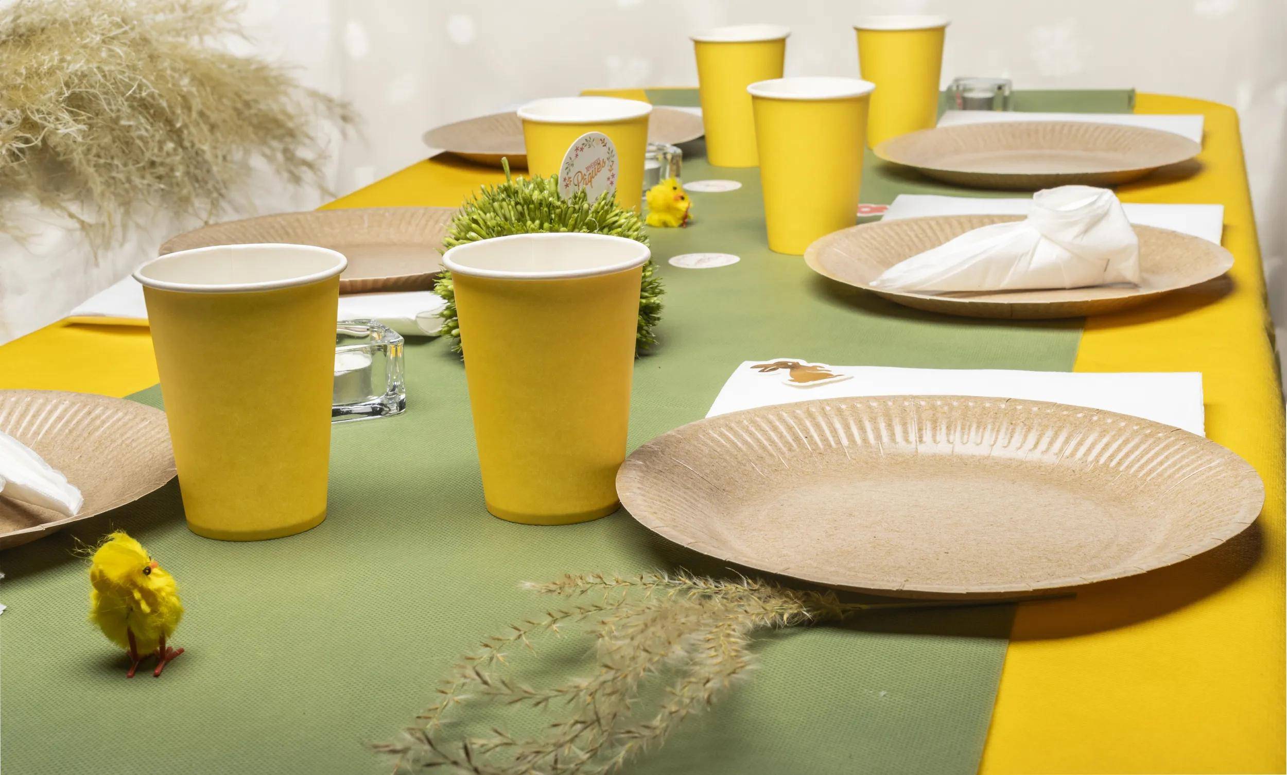 Ein grünes Tischtuch mit gelben Tassen und braunen Tellern für eine Mahlzeit eingedeckt; kleine Zimmerpflanzen und Glasgefäße sind vorhanden.
