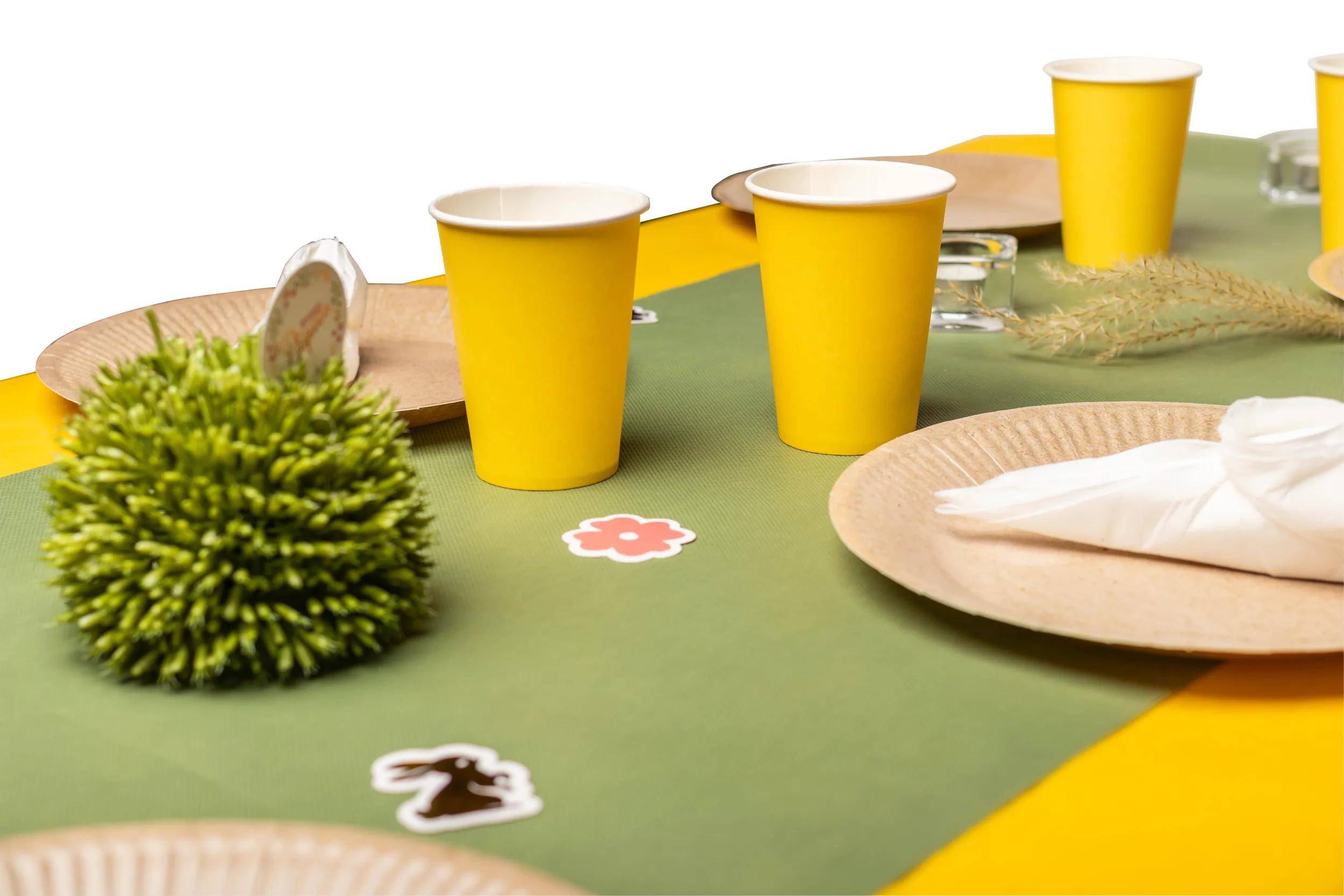 Drei gelbe Papiertassen auf einer grünen Tischdecke, umgeben von Tellern, Servietten und kleinen Dekorationen wie einer Blume und einem Häschen.