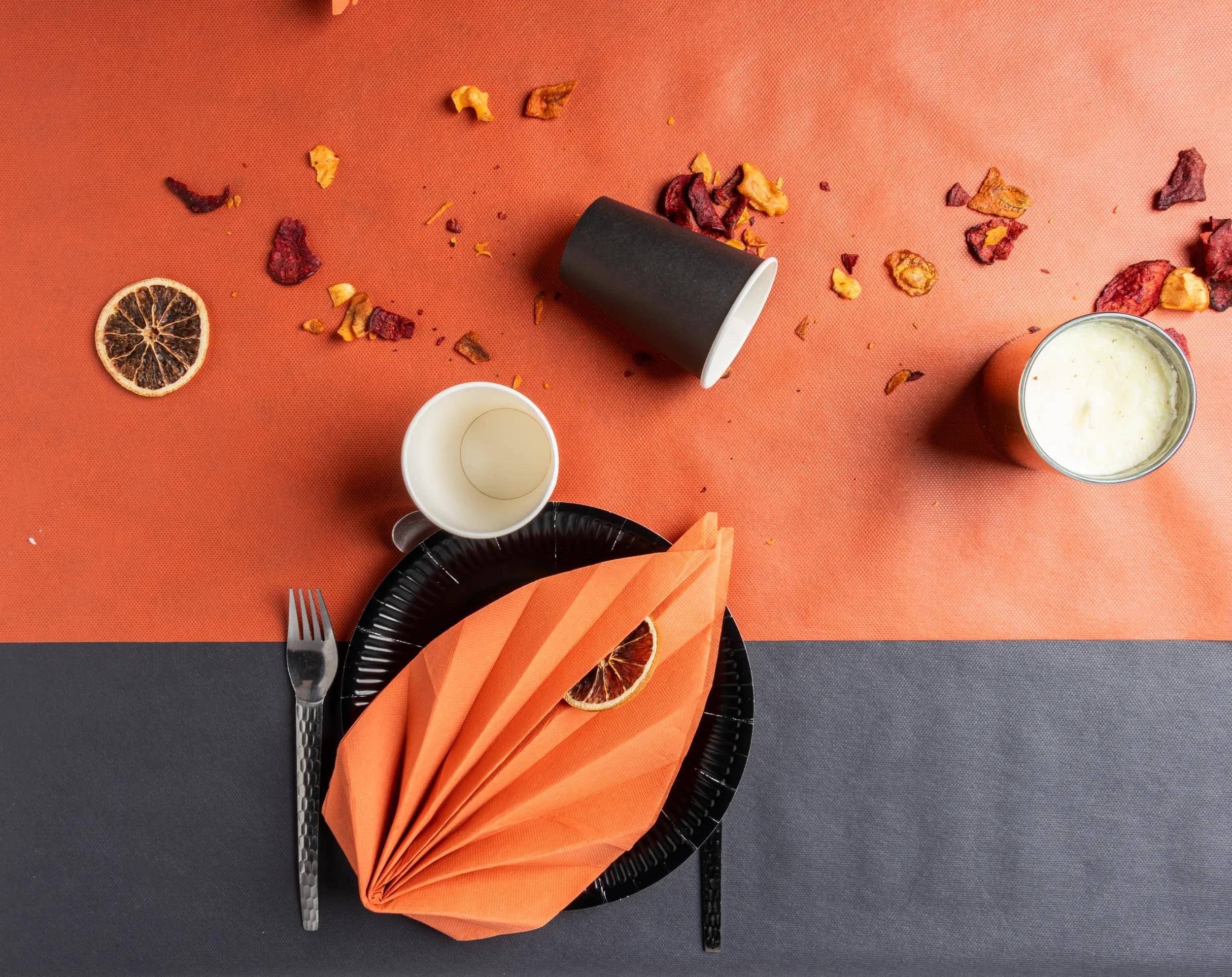 Ein Tischgedeck mit einer orange Serviette, die dekorativ auf einem schwarzen Teller gefaltet ist, begleitet von einer Gabel und einem Messer. Verstreute getrocknete Zitrusscheiben und Blütenblätter verleihen Farbe.