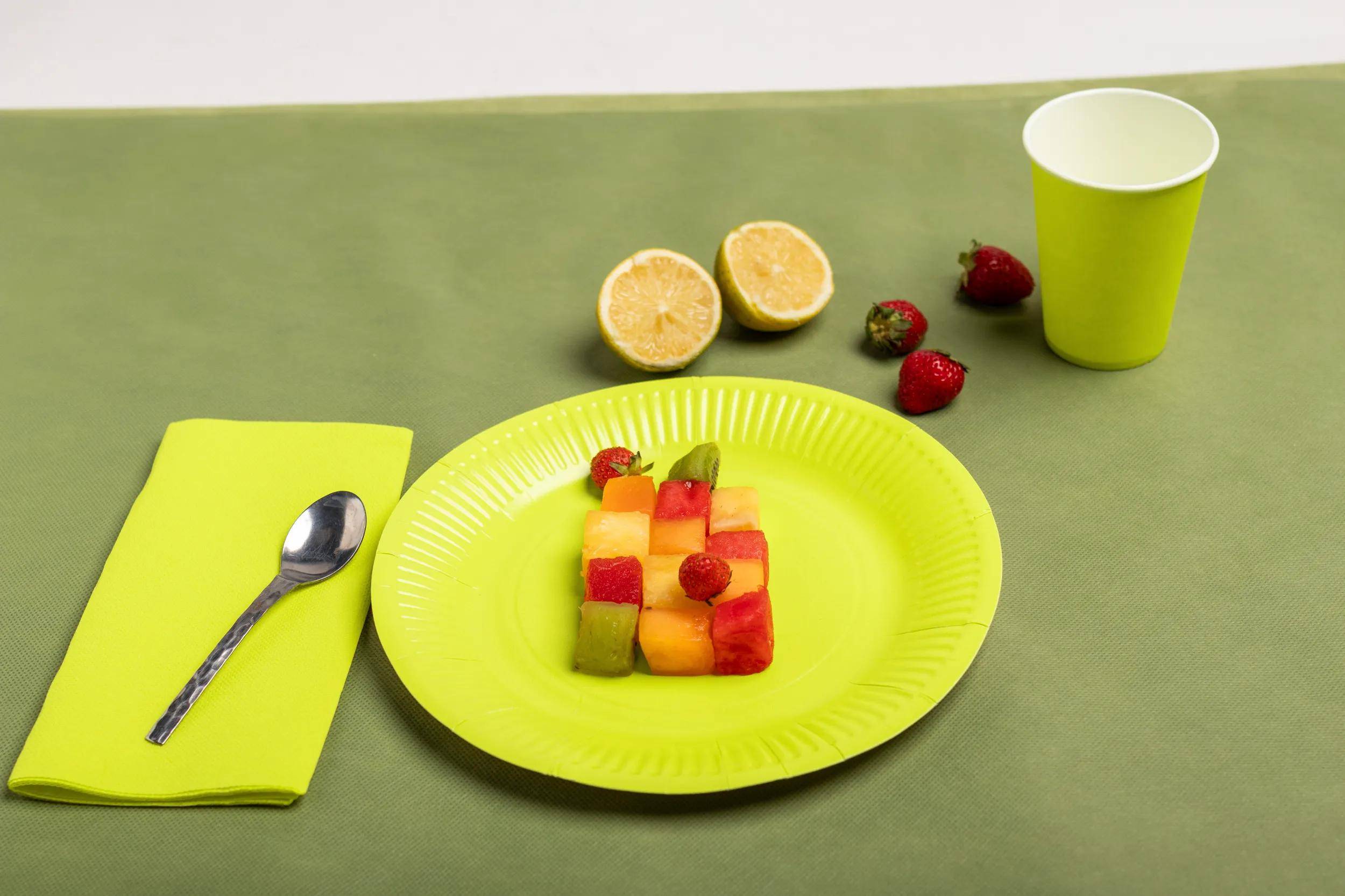 Ein Teller mit bunten Frucht­würfeln und gewürfelten Erdbeeren, daneben eine halbe Zitrone, Erdbeeren und eine Tasse auf einer grünen Platzdecke.