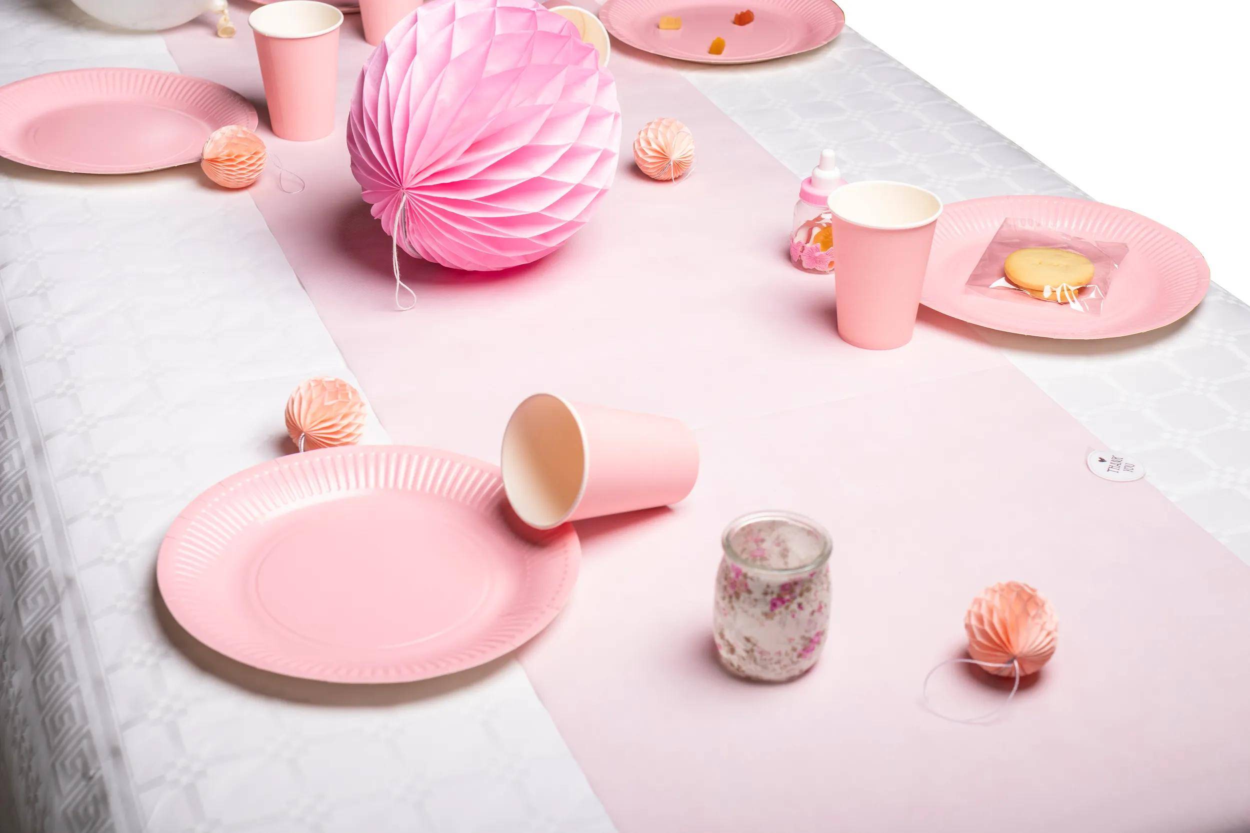 Tisch mit rosa Papiertellern, Bechern und Dekorationen, einschließlich eines großen Papierfächers und eines Blumenkerzenstänanders, was ein festliches Setup nahelegt.