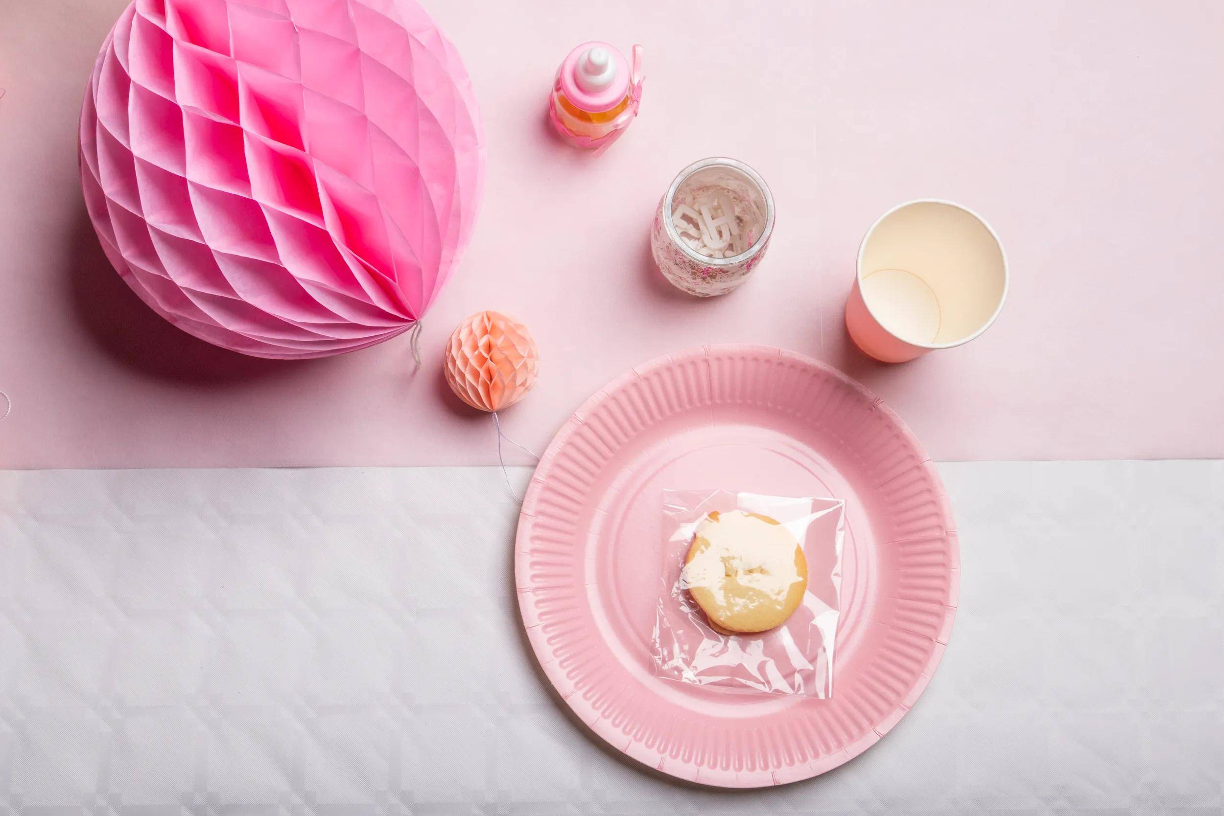 Ein rosa Papierteller mit einem eingewickelten Keks darauf sitzt auf einer rosa Tischdecke, umgeben von dekorativen Papierkugeln und Tassen.
