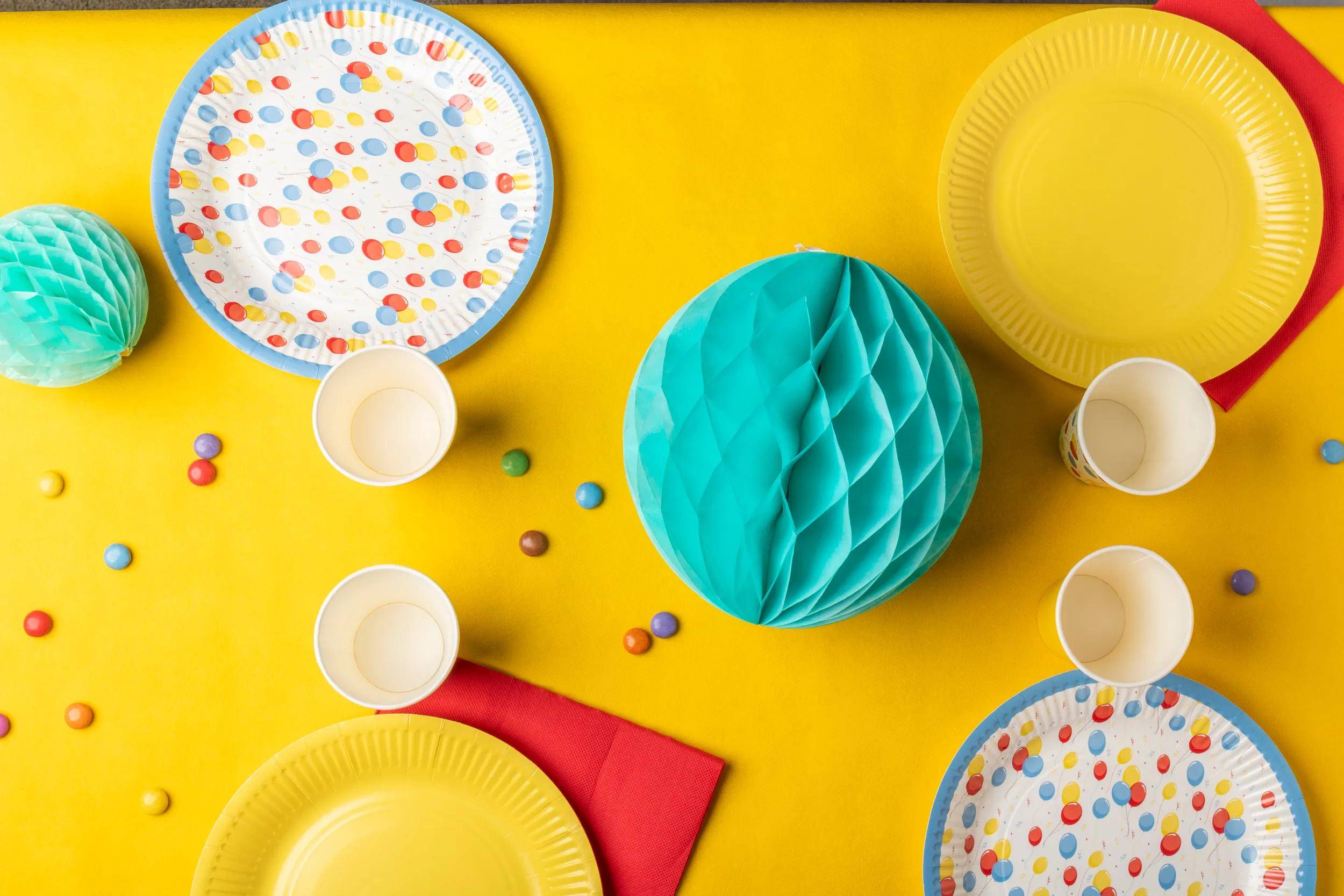Farbenfrohe Tischdecke mit Tellern im Polka-Dot-Muster, gelben Tellern, roter Serviette, Tassen und einer türkisfarbenen Tischdekoration auf einem hellen gelben Hintergrund.