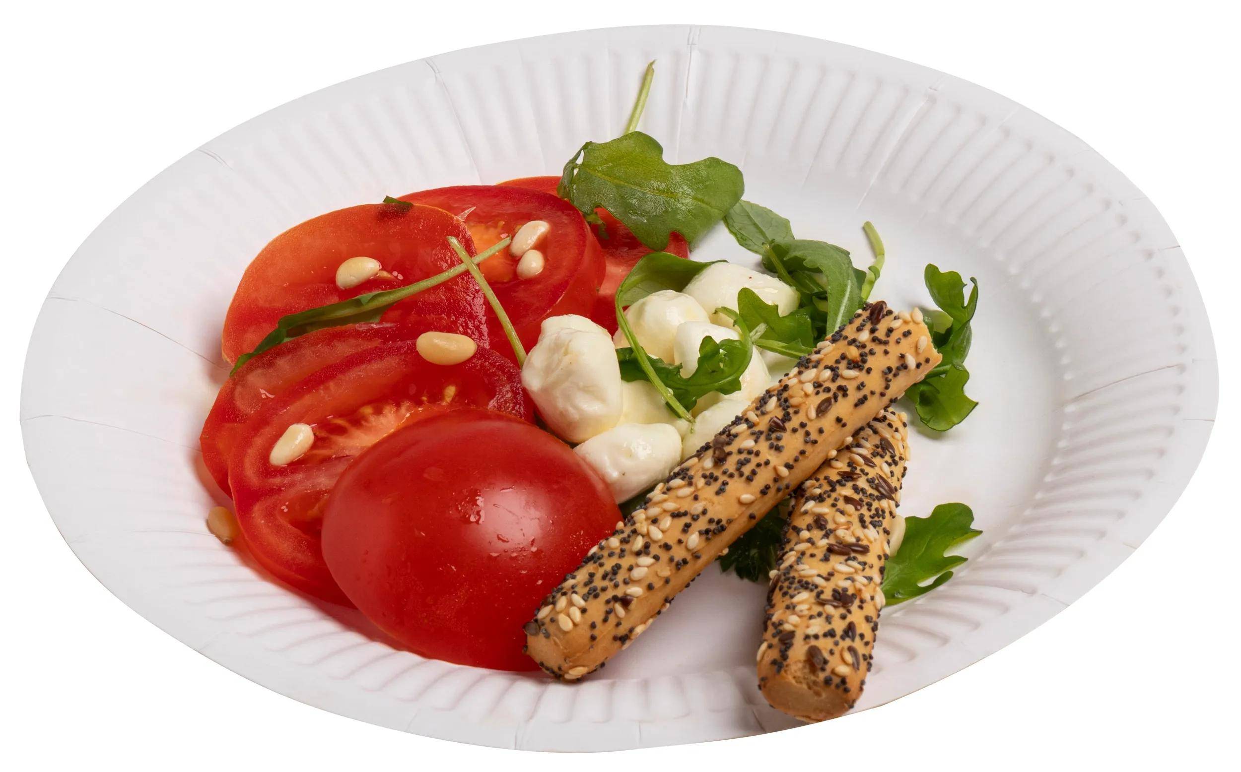 Ein weißer Papperteller zeigt einen Salat mit frischen Tomatenscheiben, Mozzarellakugeln, Rucola, Pinienkernen und Sesamstangen.