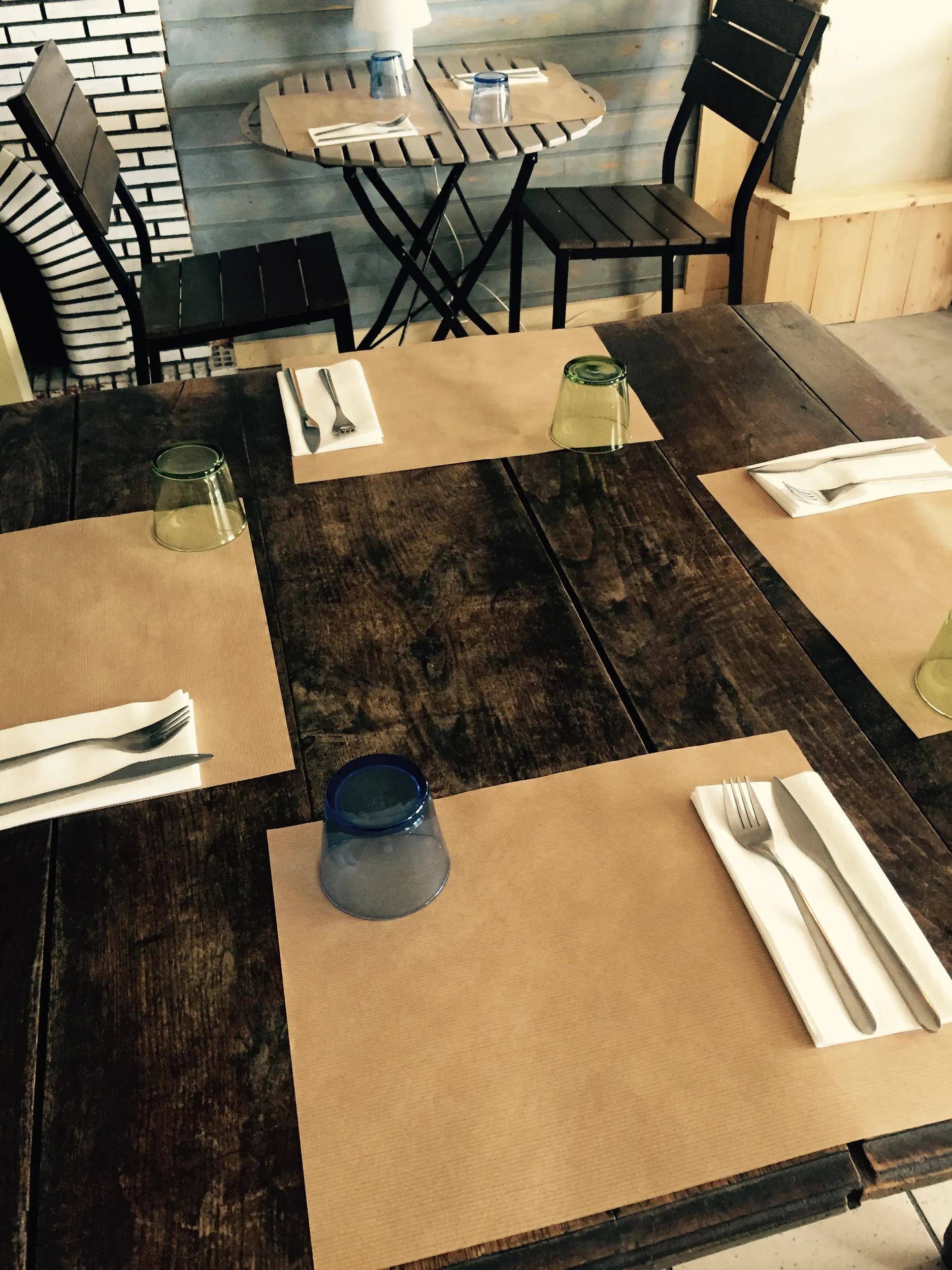 Ein Holztisch ist mit braunen Platzdeckchen, weißen Servietten und Besteck gedeckt. Klare Gläser sind bei jedem Gedeck umgestülpt platziert.