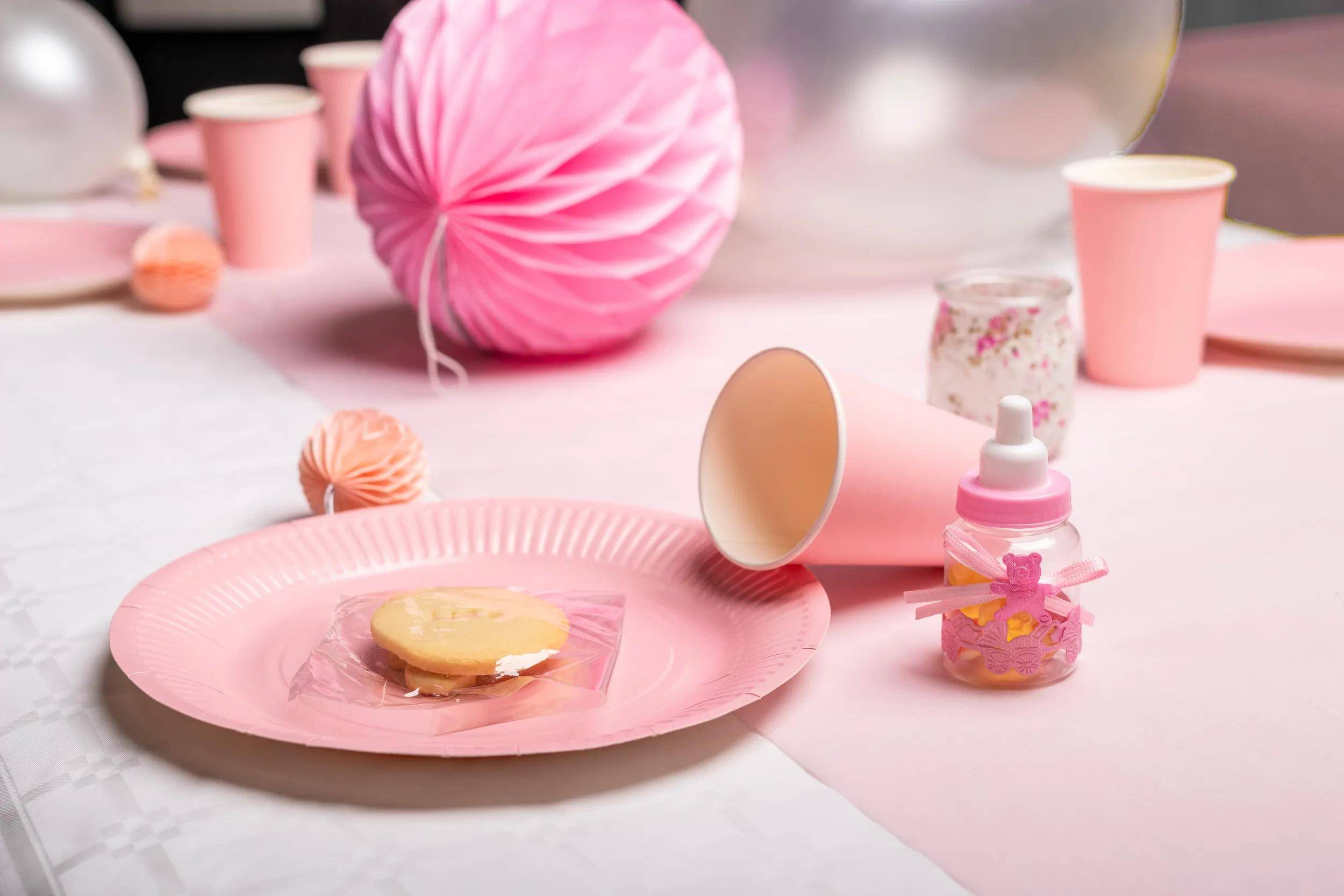 Pink-Party-Setup mit einem herzförmigen Keks auf einem Teller, einer Babyflasche mit rosa Schleife und einem Pappbecher, der auf der Seite liegt.