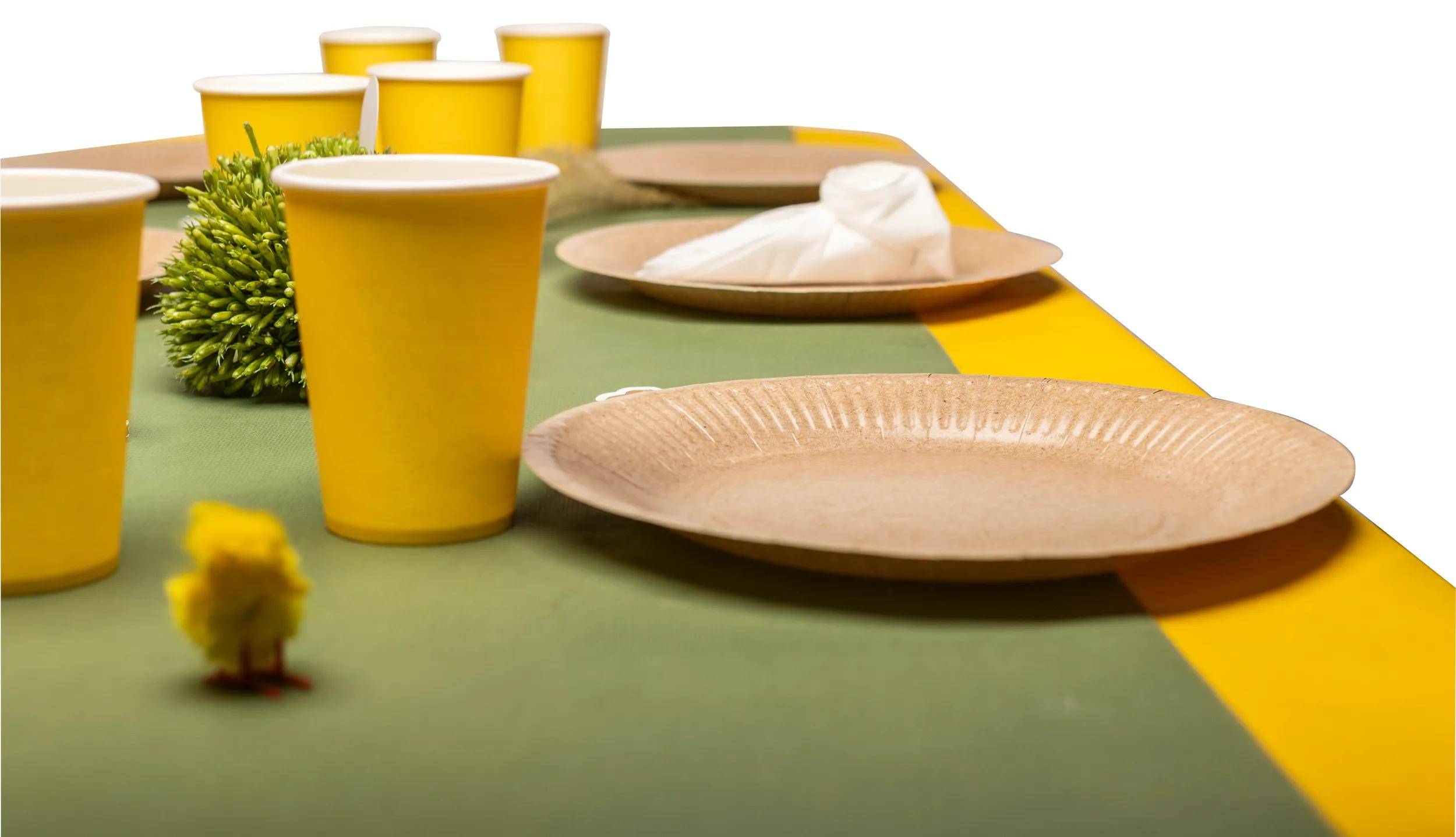 Gelbe Papiertassen und biologisch abbaubare Teller auf einem grünen Tisch mit einem gelben Tischläufer, was ein umweltfreundliches Versammlungssetting andeutet.