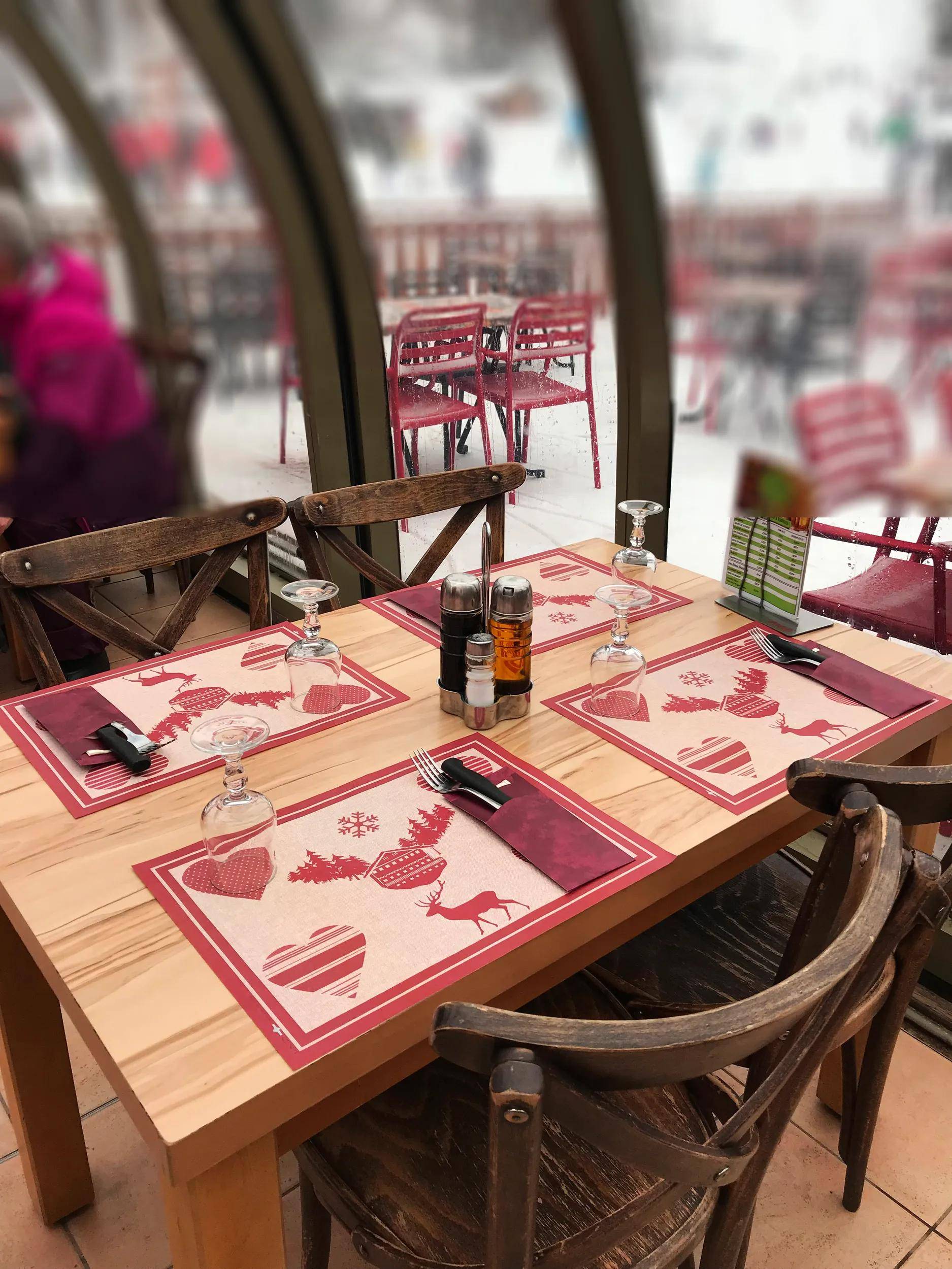 Ein gemütlich gedeckter Restauranttisch für vier Personen mit herzförmigen Platzsets, Besteck, Gewürzen und einem Blick auf einen schneebedeckten Außenbereich.