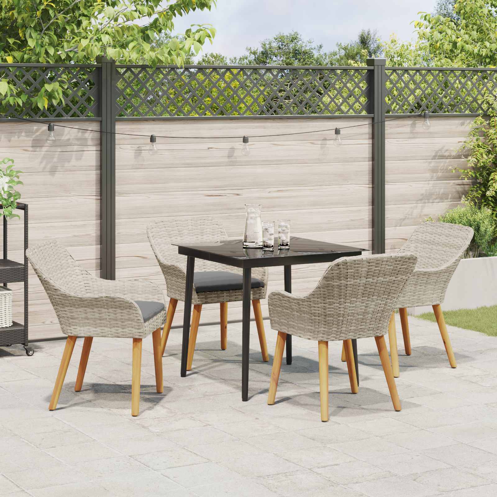 Ein gemütlicher Außenpatio mit einem kleinen schwarzen Tisch und vier Korbstühlen vor einem Sichtschutzzaun, umgeben von üppiger Bepflanzung.