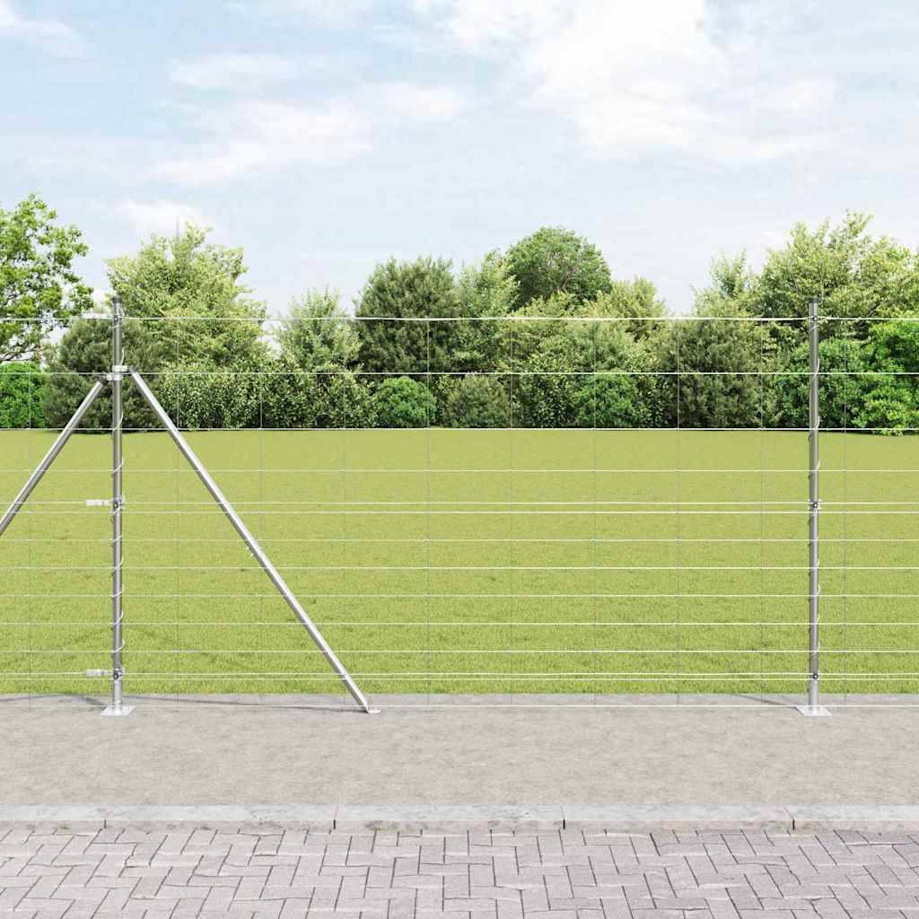 Ein Maschendrahtzaun steht auf Beton, umgeben von Gras und üppig grünen Bäumen, unter einem klaren blauen Himmel.