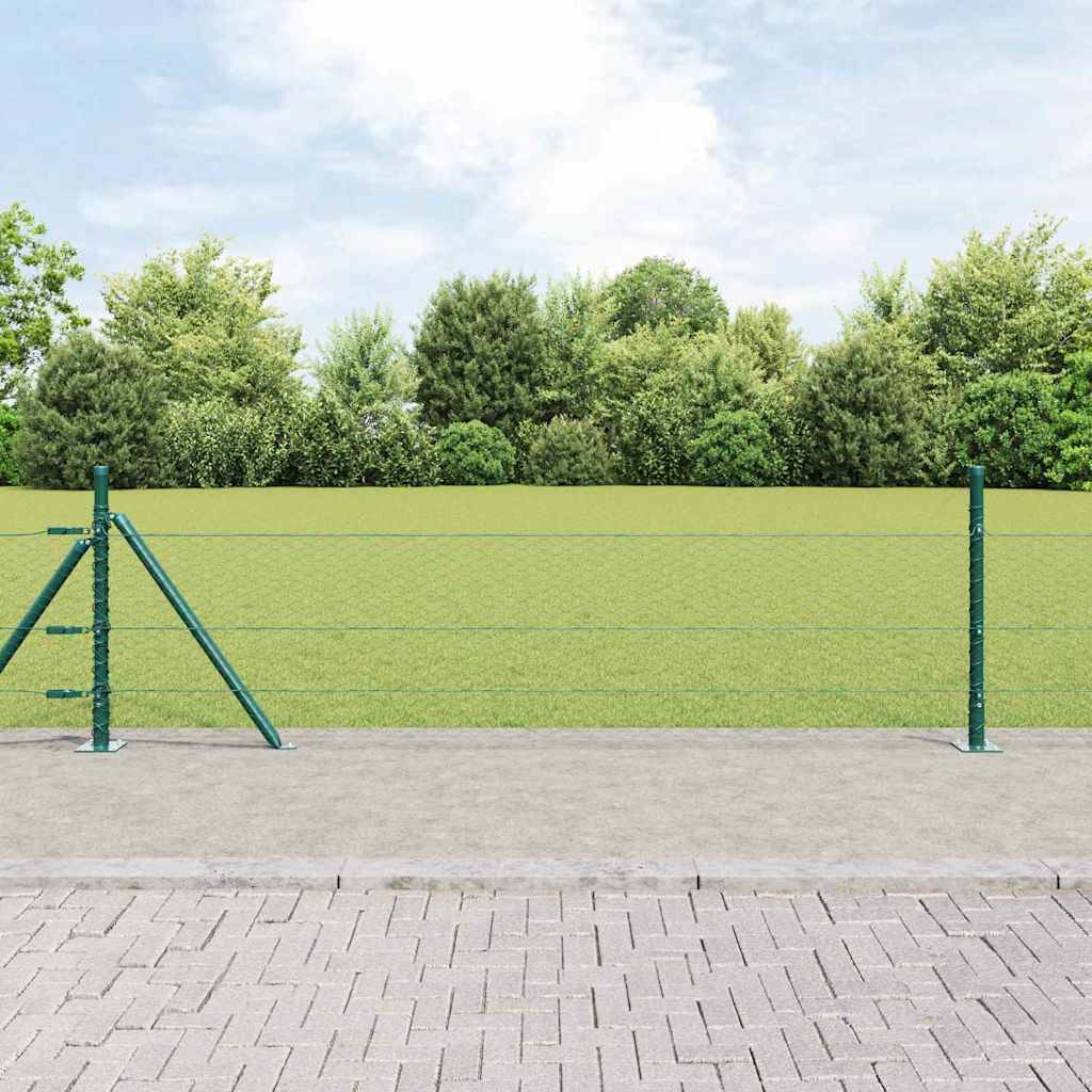 Ein Stacheldrahtzaun trennt eine gepflasterte Fläche von einem grasbewachsenen Feld mit dichten Bäumen im Hintergrund unter einem bewölkten Himmel.